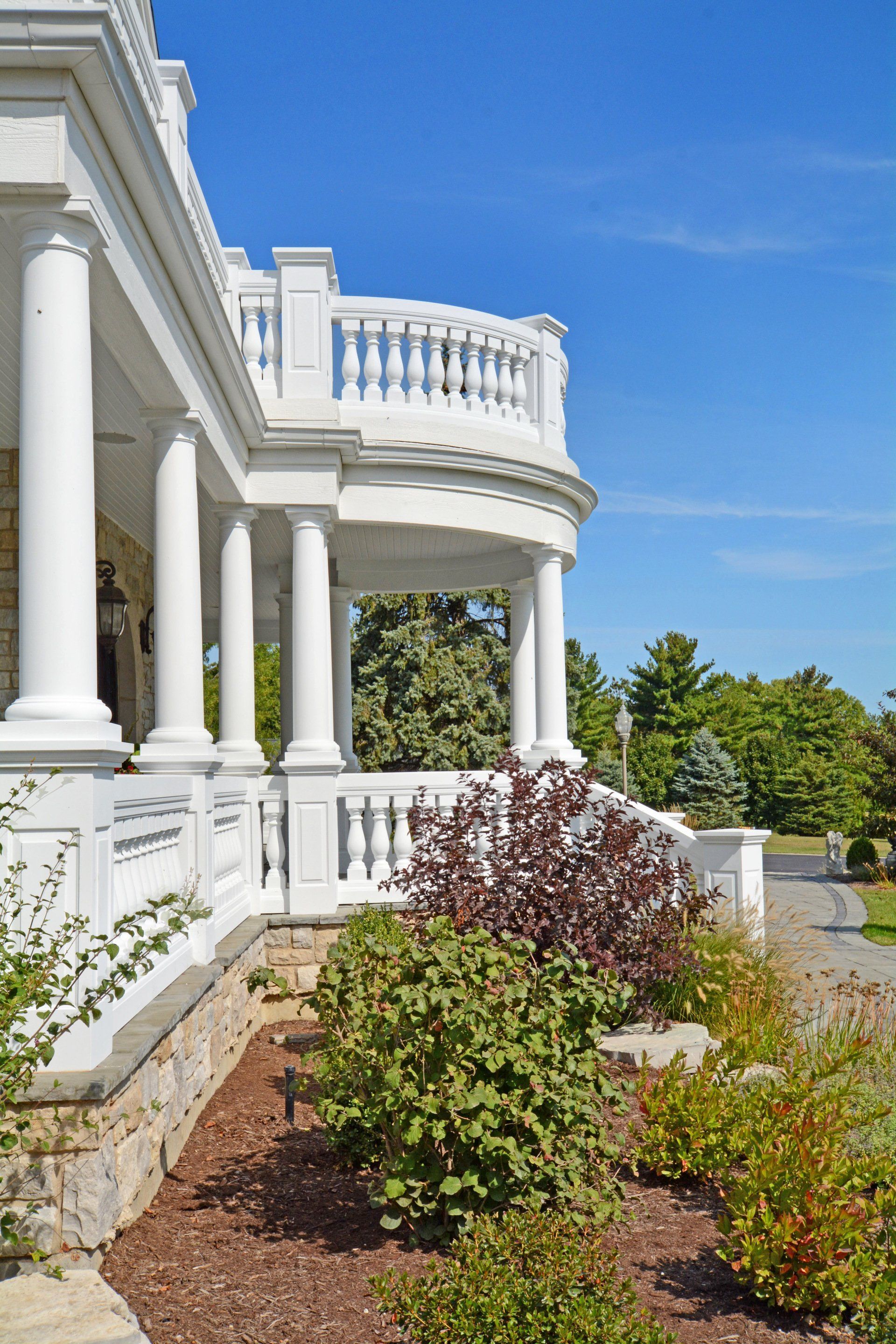 White columned porch with balcony, overlooking a landscaped garden under a blue sky.