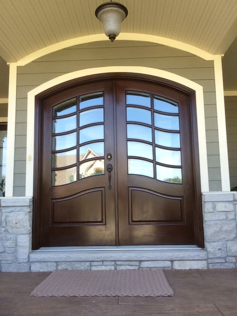 Double wooden front door with arched glass panes and a doormat under a porch light