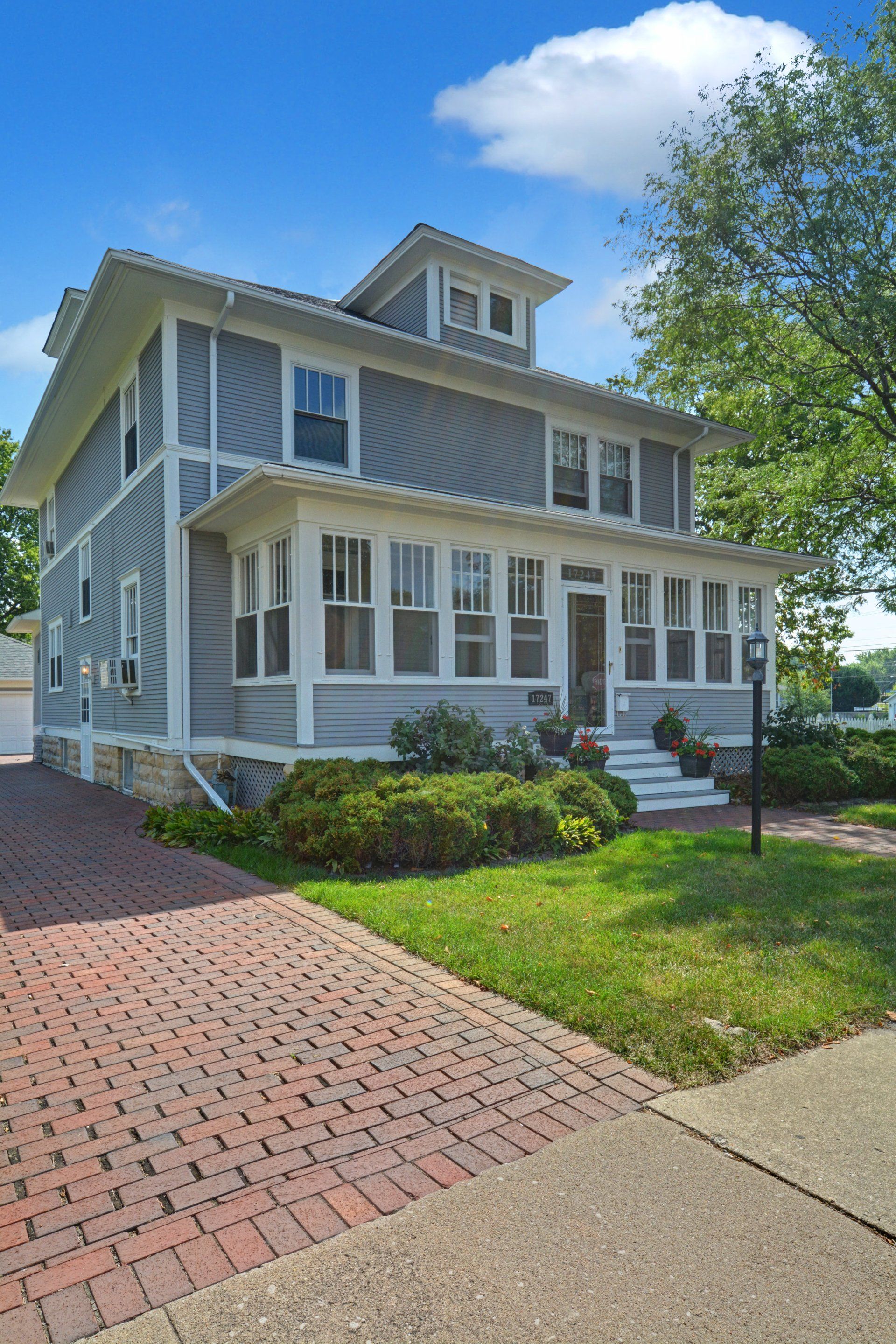 White two-story house with porches, brick driveway, and green lawn on a sunny day