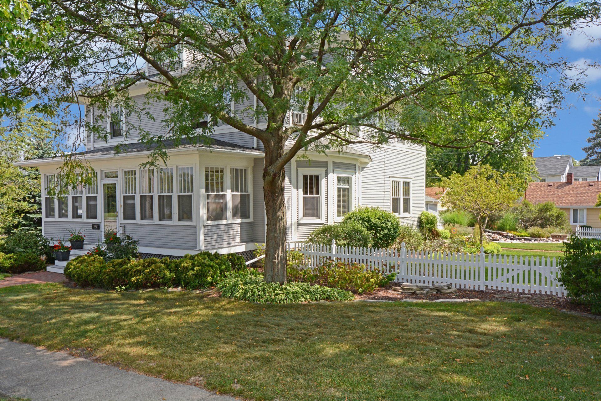 White house with porch and picket fence beside a tree on a sunny residential street