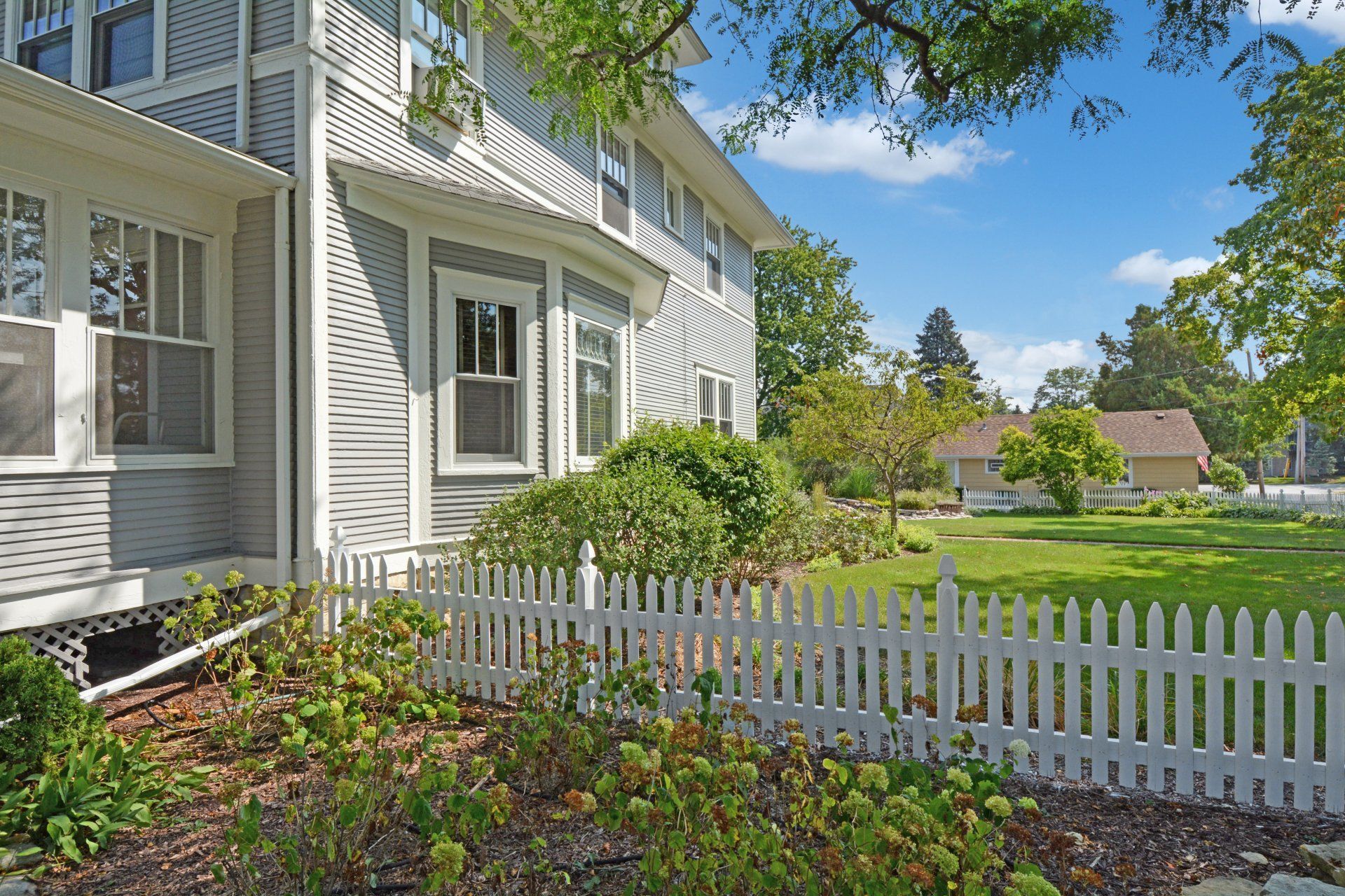 White house with a white picket fence, green lawn, and garden under a blue sky
