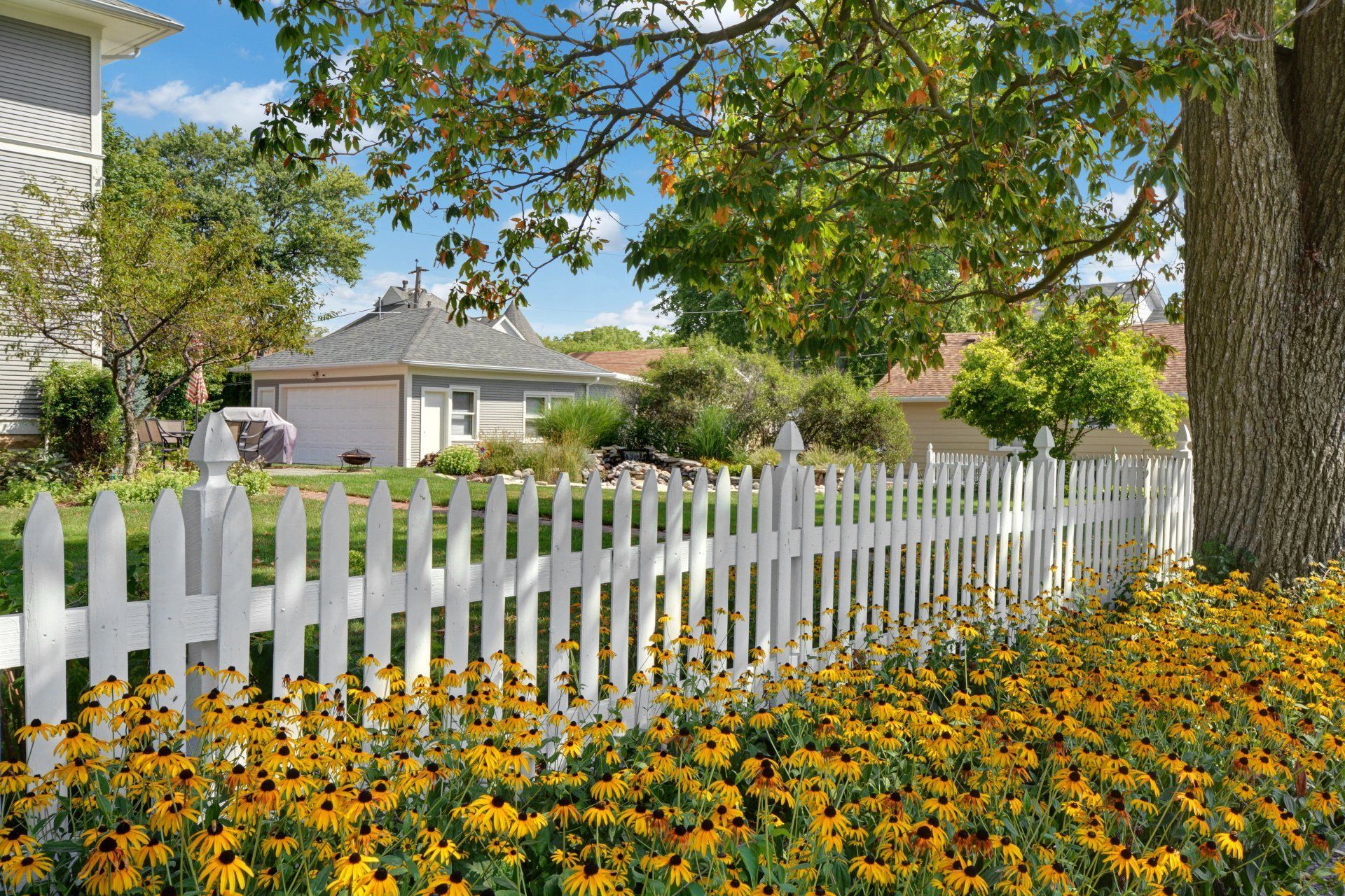 White picket fence beside yellow flowers and a house under leafy trees on a sunny day