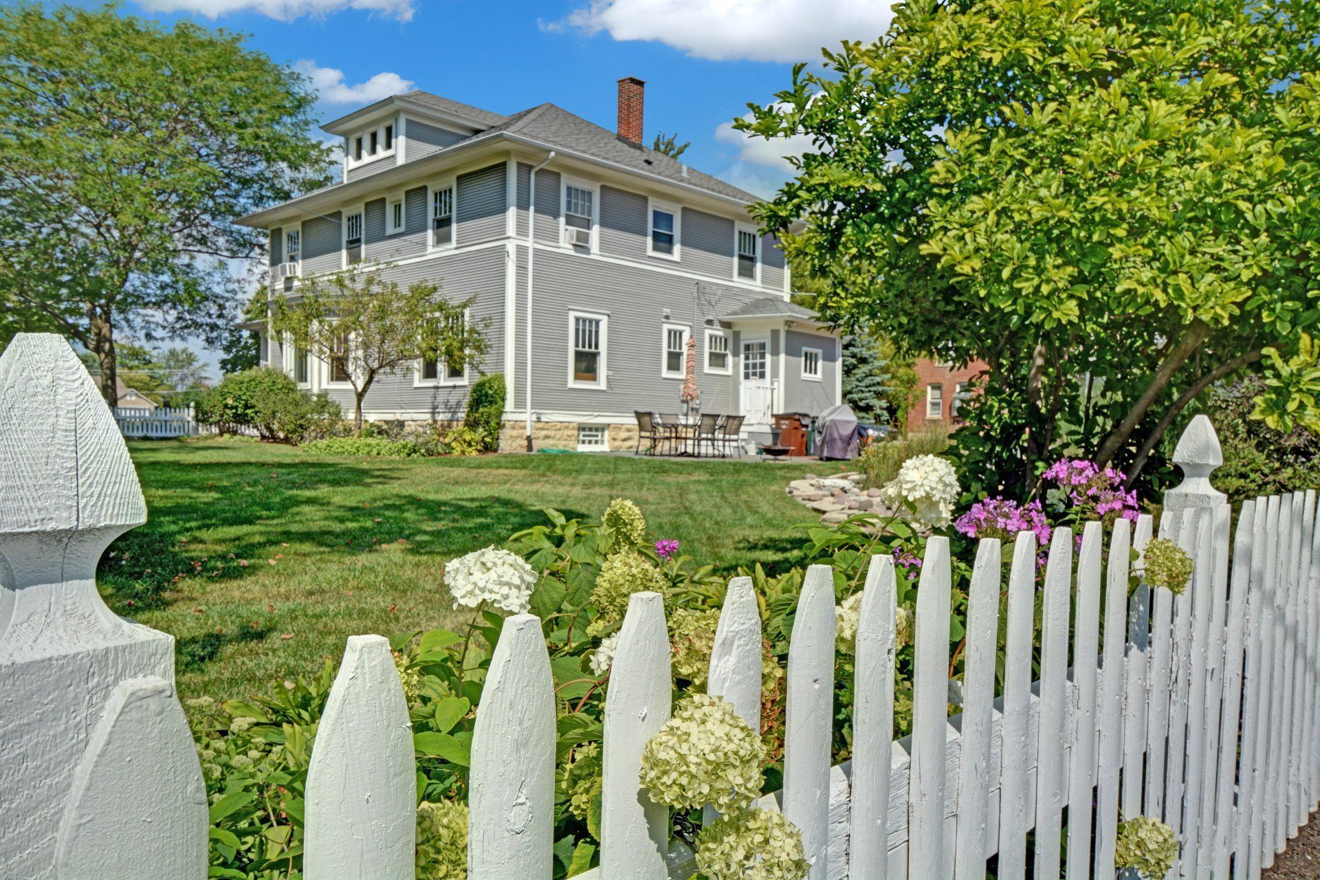 White picket fence in front of a gray house with a green lawn and blooming garden under a clear blue sky