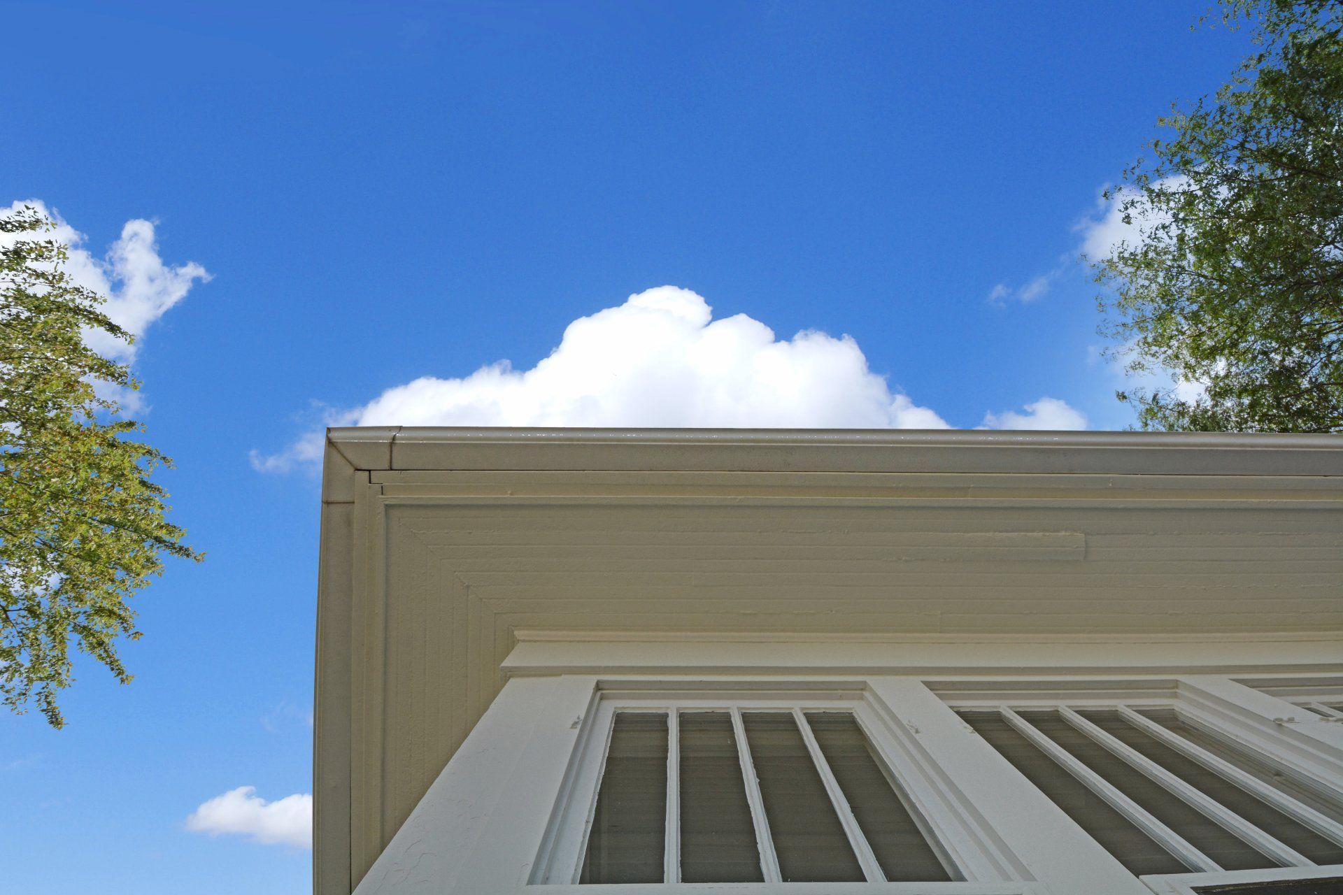 House eaves and white windows against a bright blue sky with clouds and trees