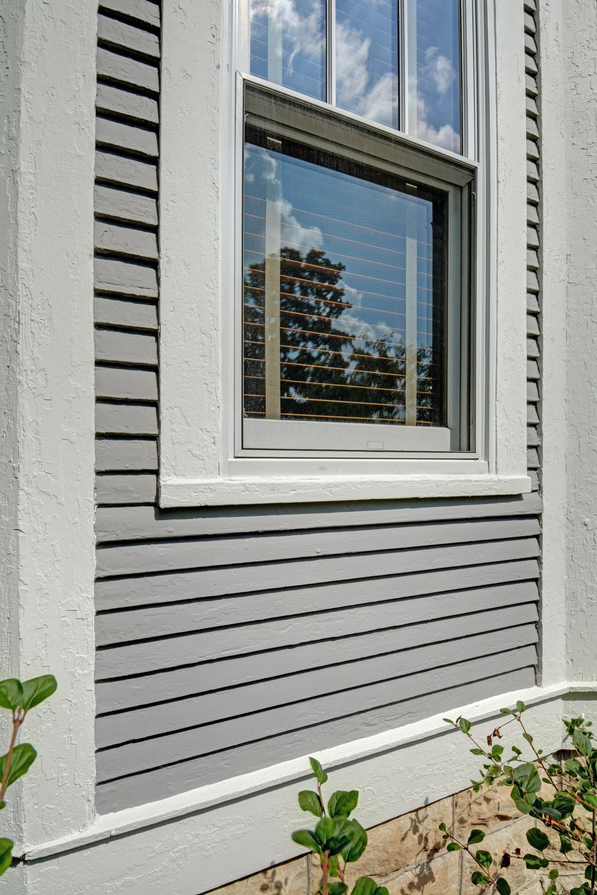 Gray house siding with a white-trimmed window reflecting trees and sky