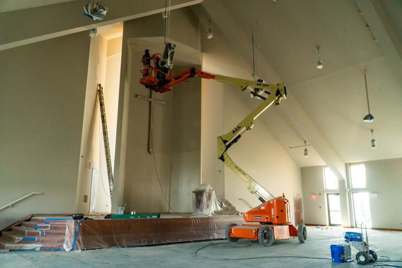 Construction workers using a lift to paint a tall wall inside a bright unfinished room