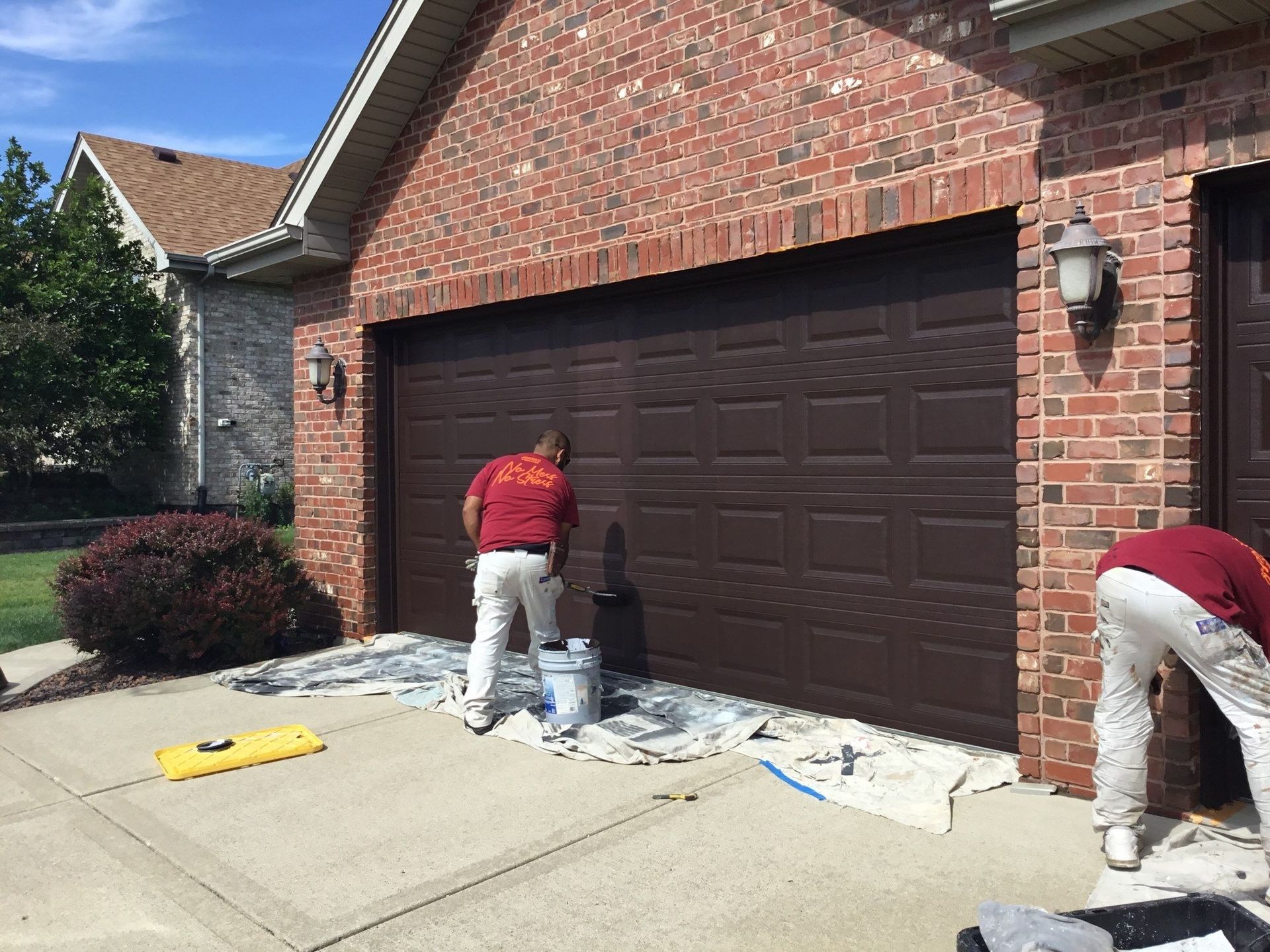 Two workers repainting a brick garage door in front of a house