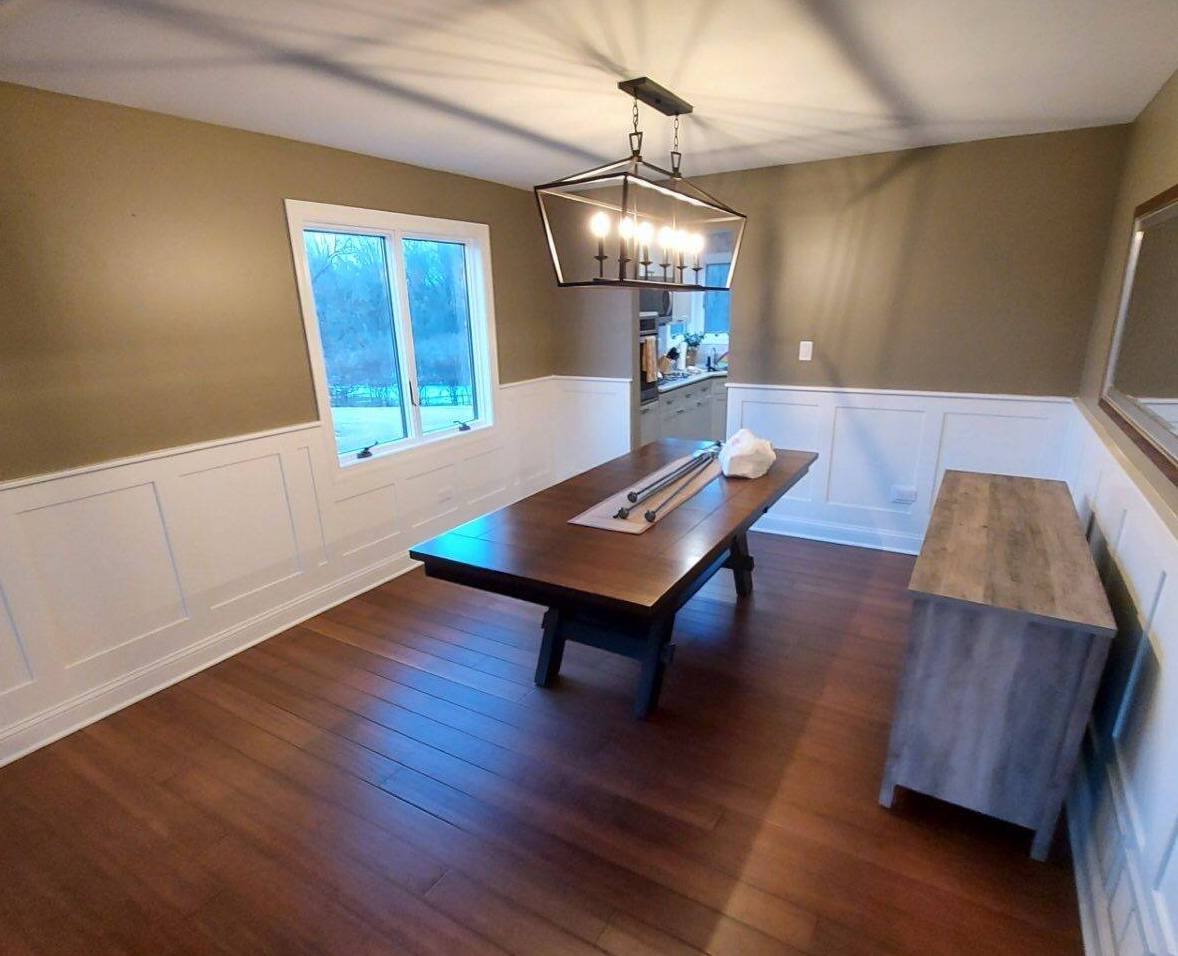 Dining room with long wooden table, chandelier, hardwood floors, and white wainscoting by a window.