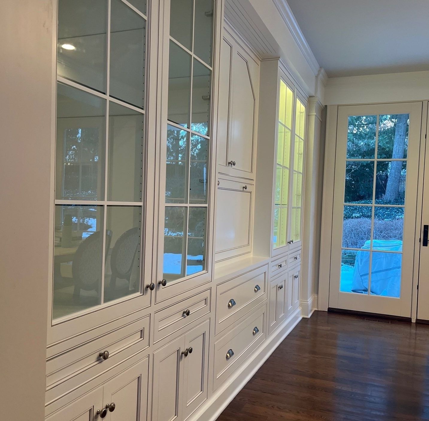 White built-in cabinets with glass doors in a bright hallway, leading to a blue-lit room and outdoor view.