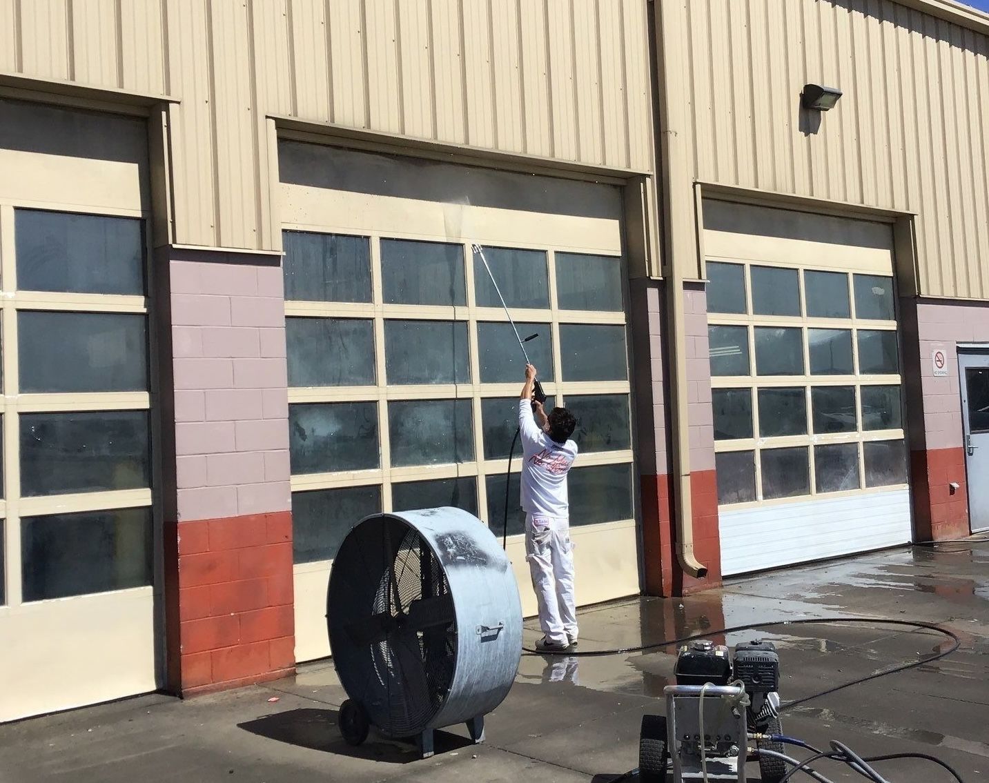 Person in white work clothes standing by a large industrial fan outside a beige garage building.