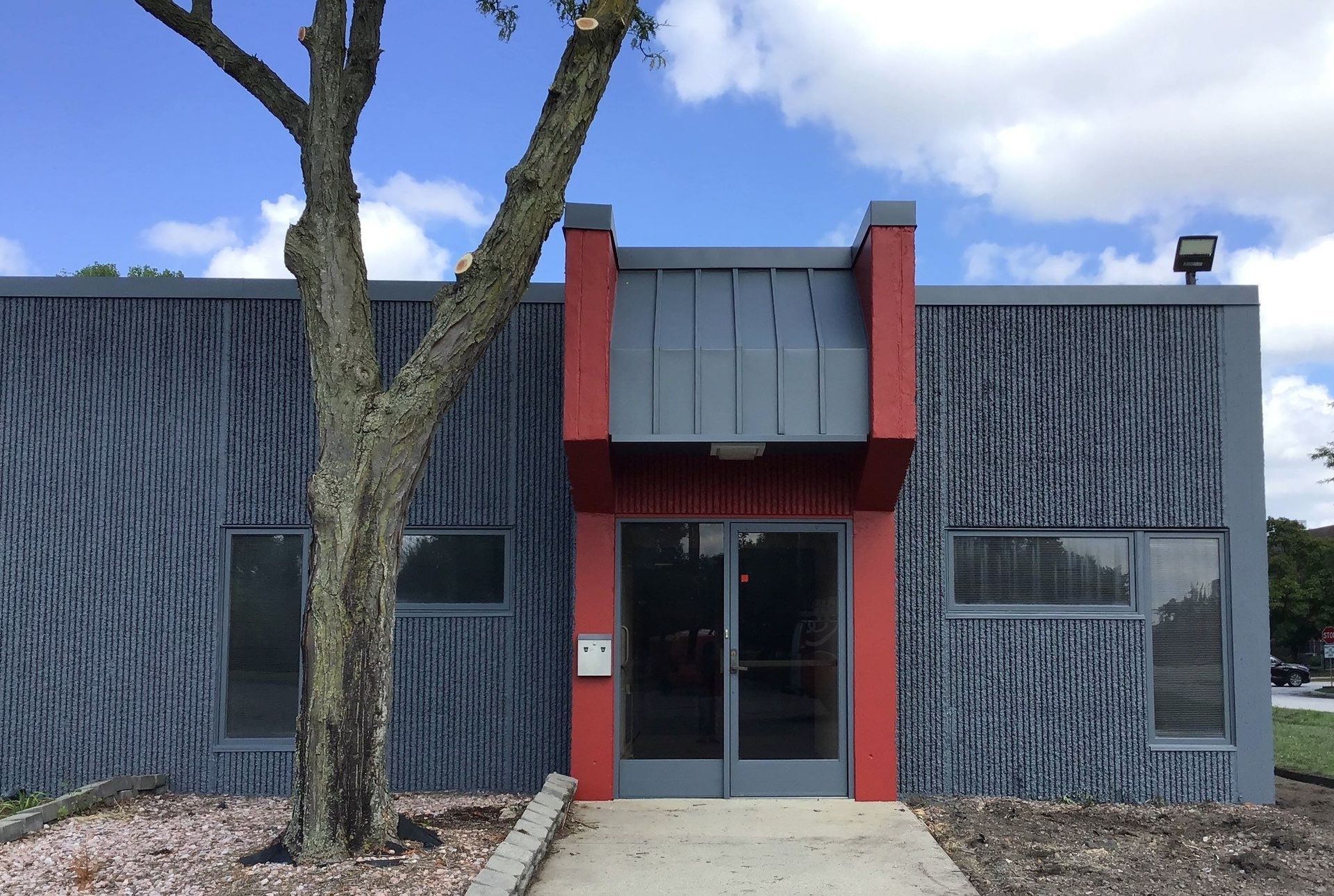 Modern building entrance with red trim, gray siding, two windows, and a tree in front