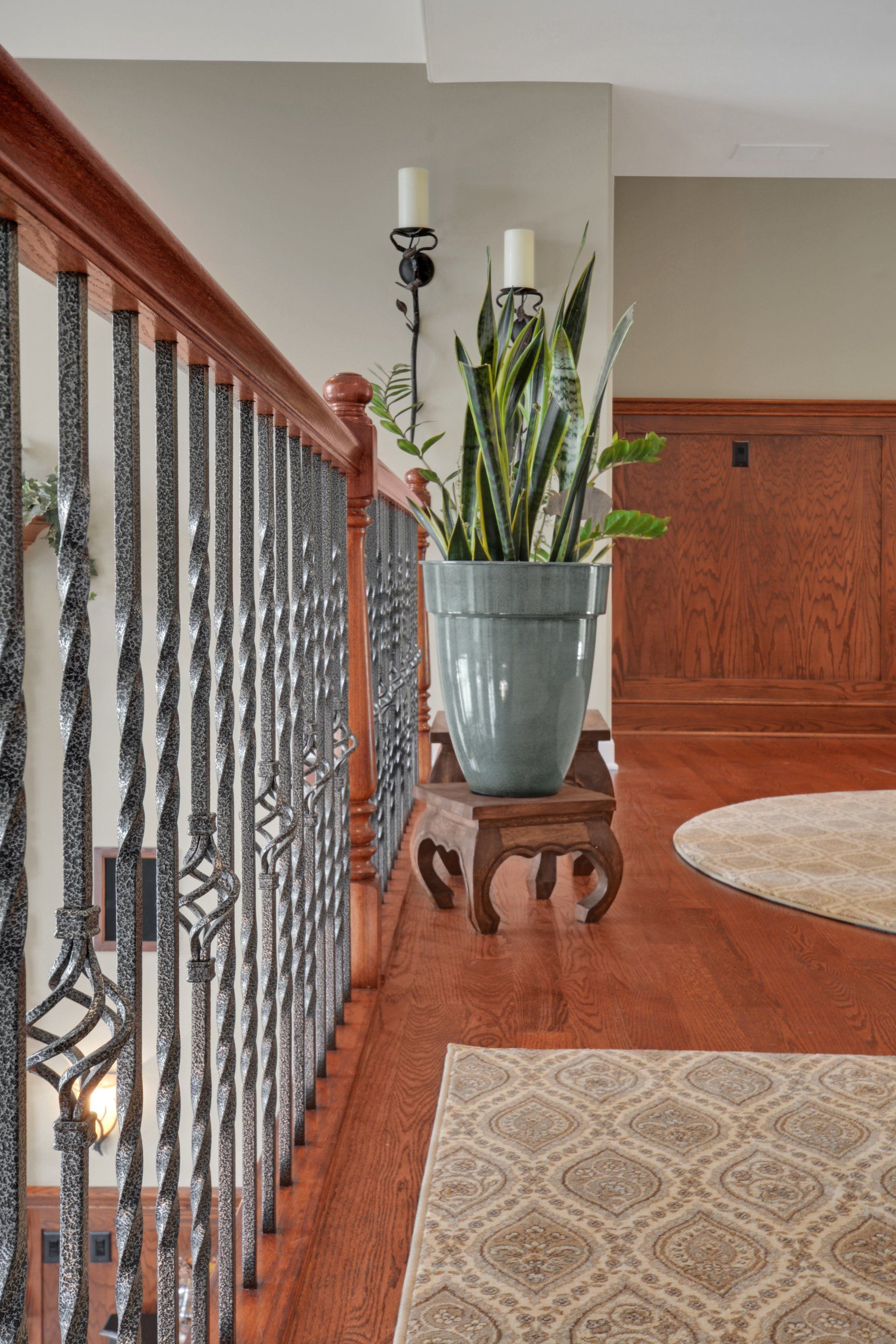 Hallway with a tall potted plant on a wooden stand beside a black metal stair railing and patterned rugs