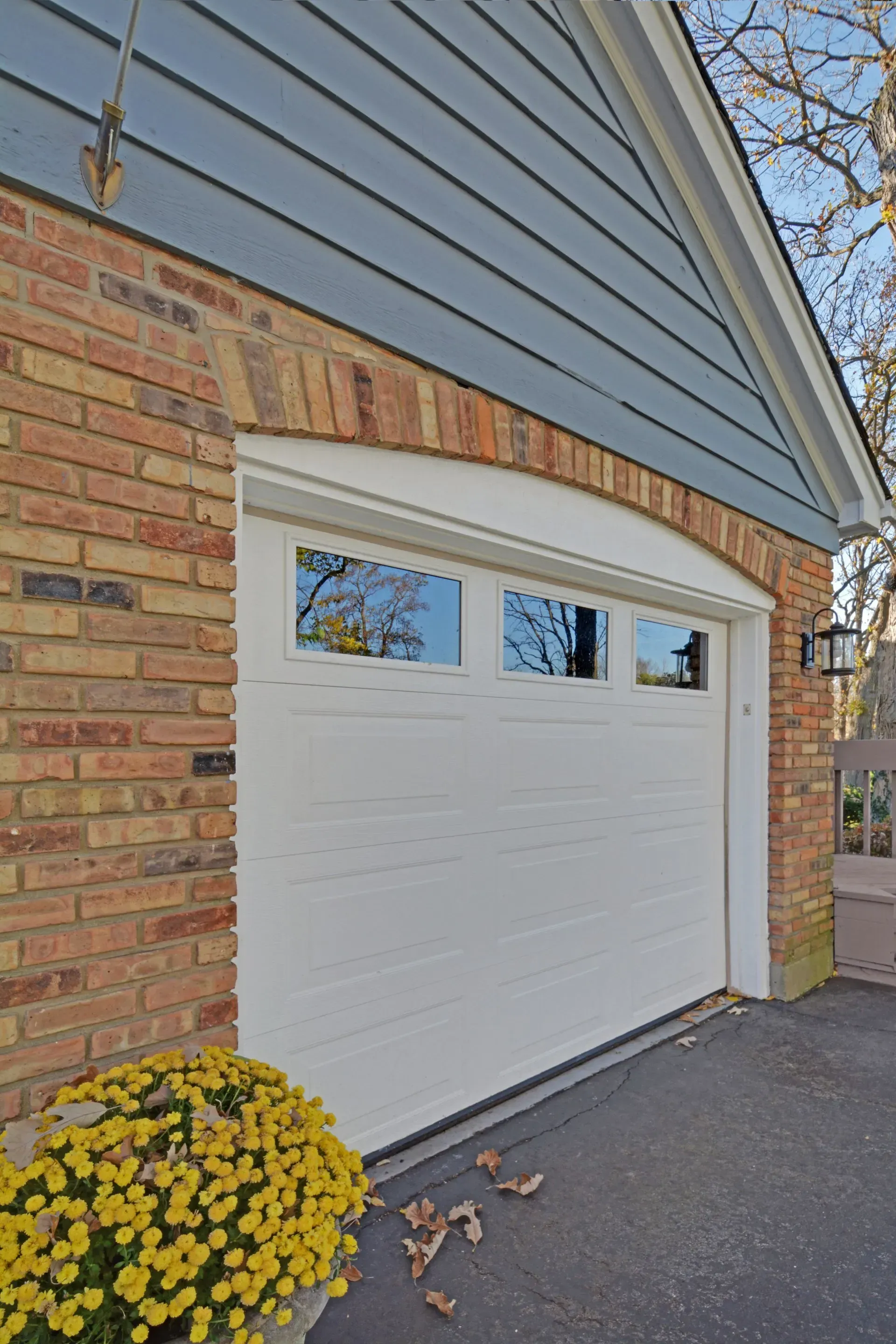 White garage door on a brick-and-blue house with a small yellow flower bush nearby.