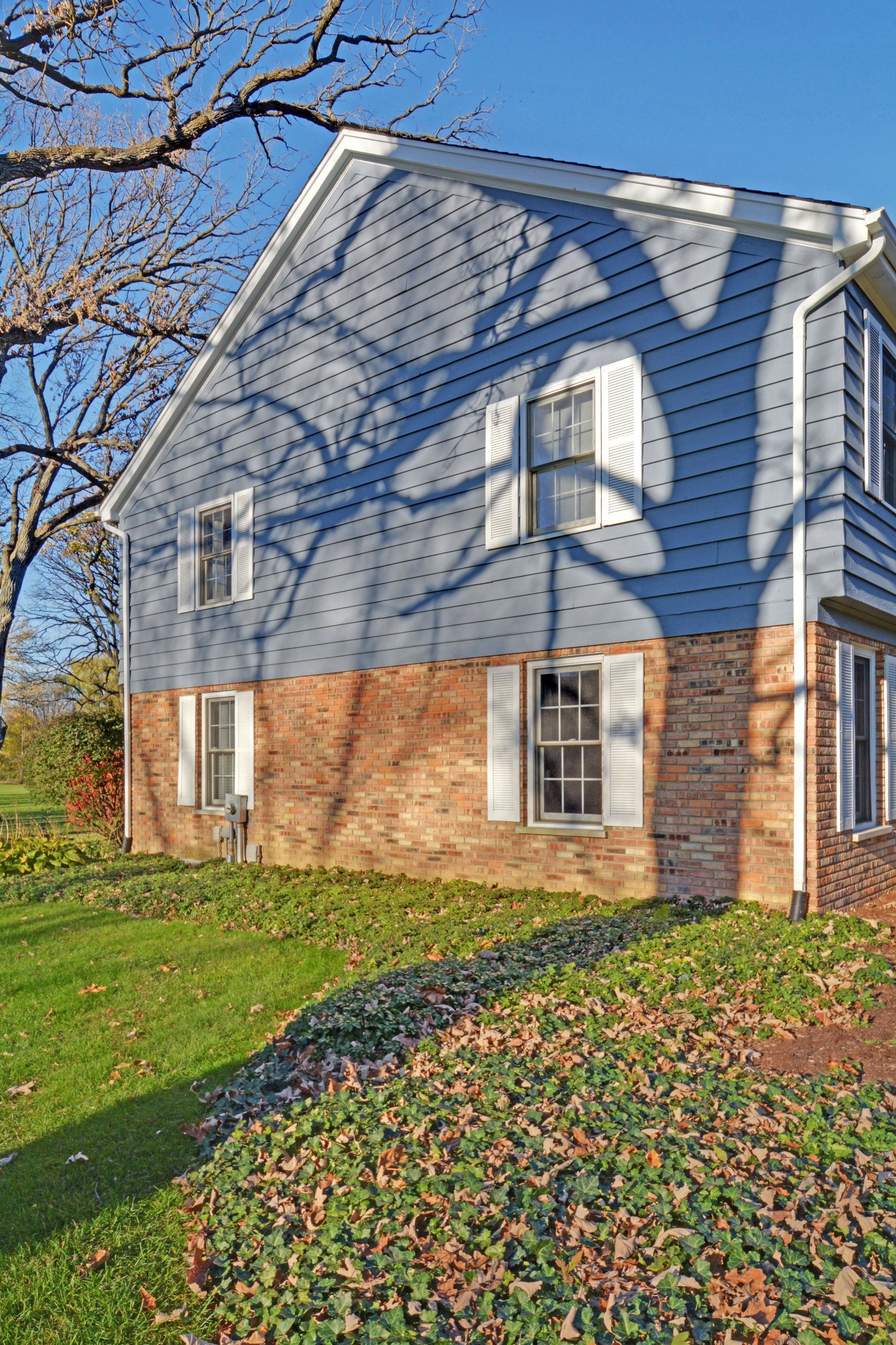 Blue-sided house with red brick base on a sunny lawn, seen beside leafless trees and a blue sky.