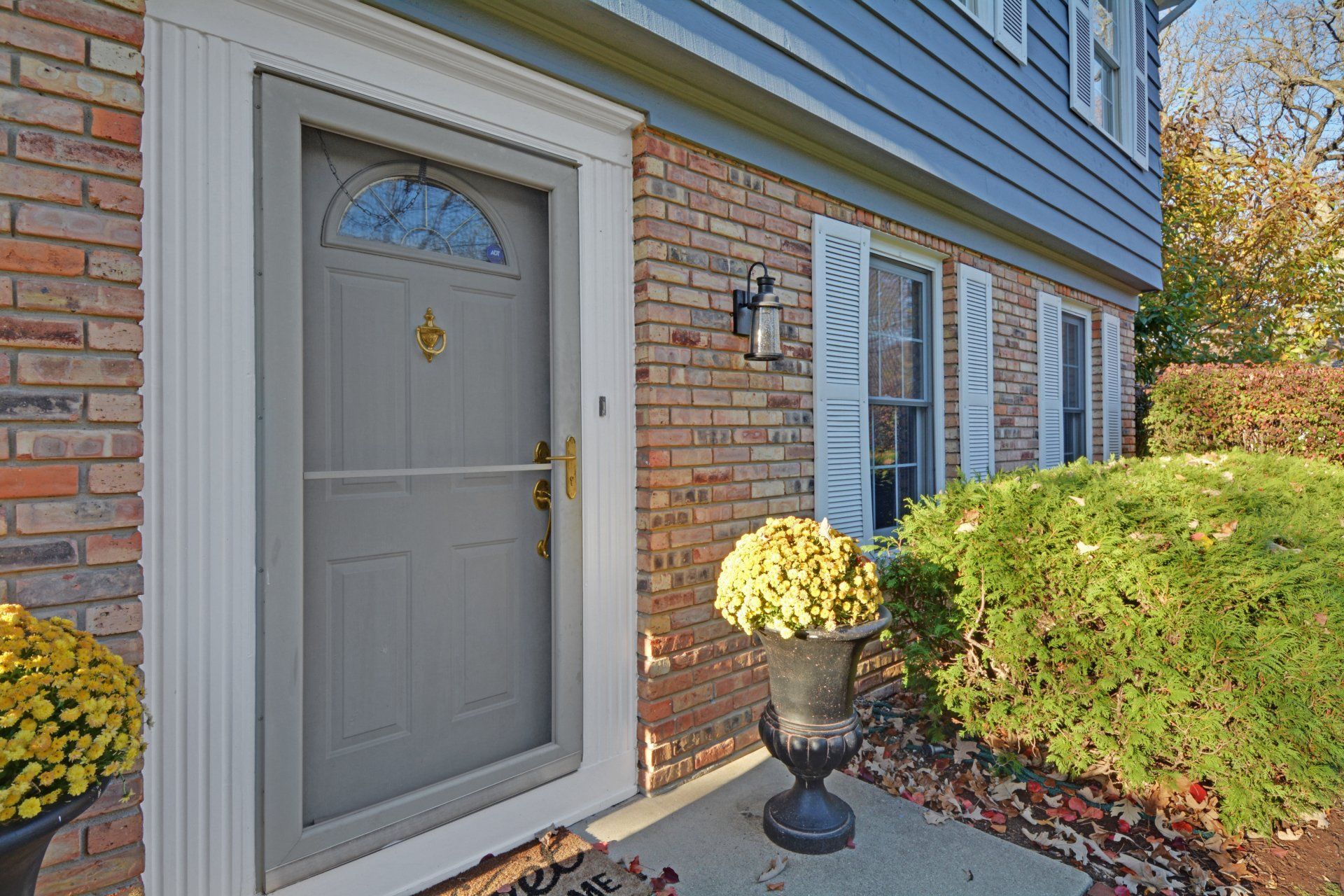 Front entry of a brick townhouse with a gray door, blue siding, and potted yellow flowers beside trimmed bushes.