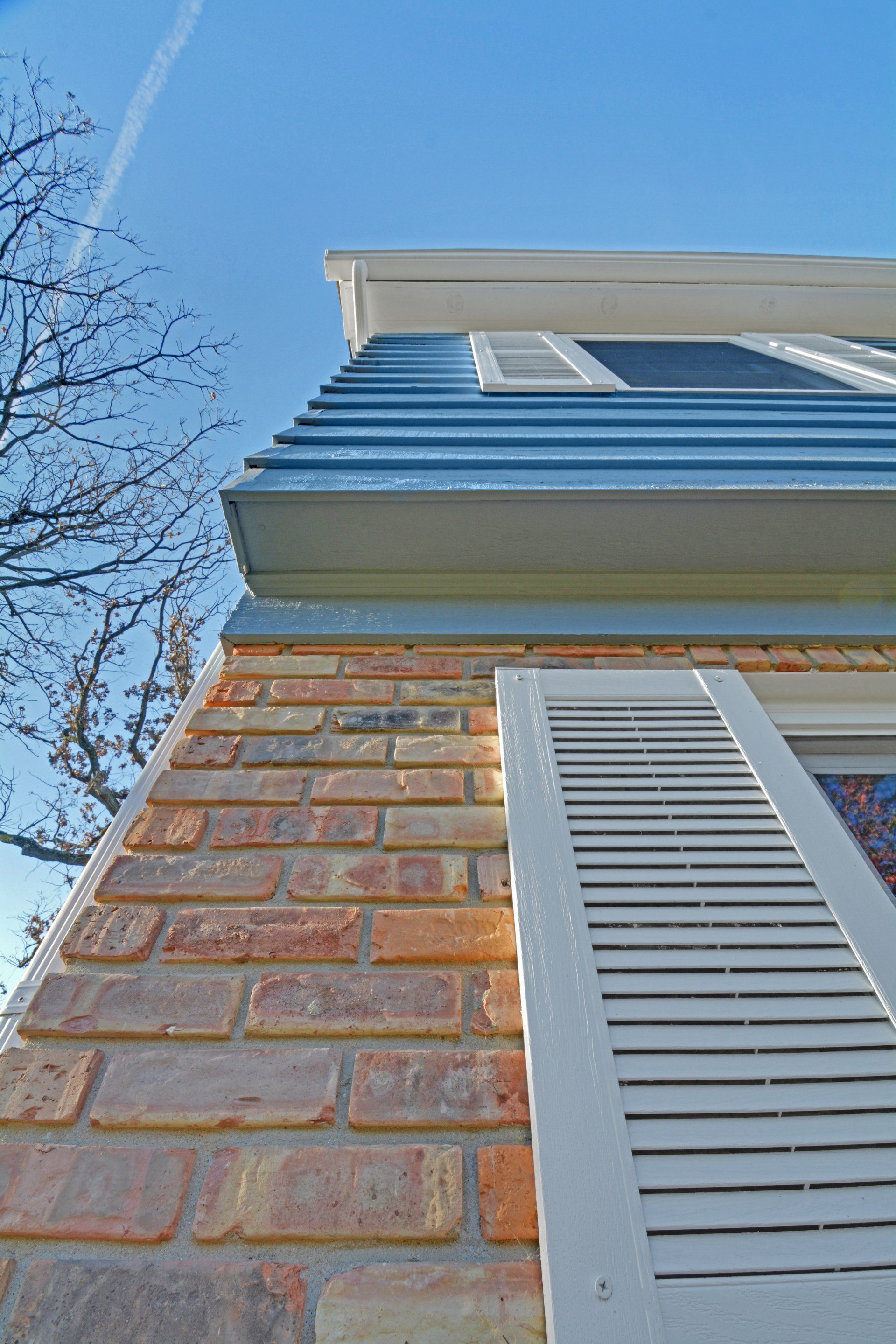 Upward view of a brick house exterior with white shutters and blue siding against a clear sky
