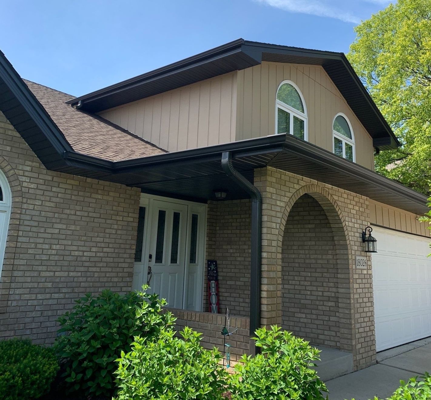 Brick house exterior with arched porch, white garage door, and green shrubs under a blue sky