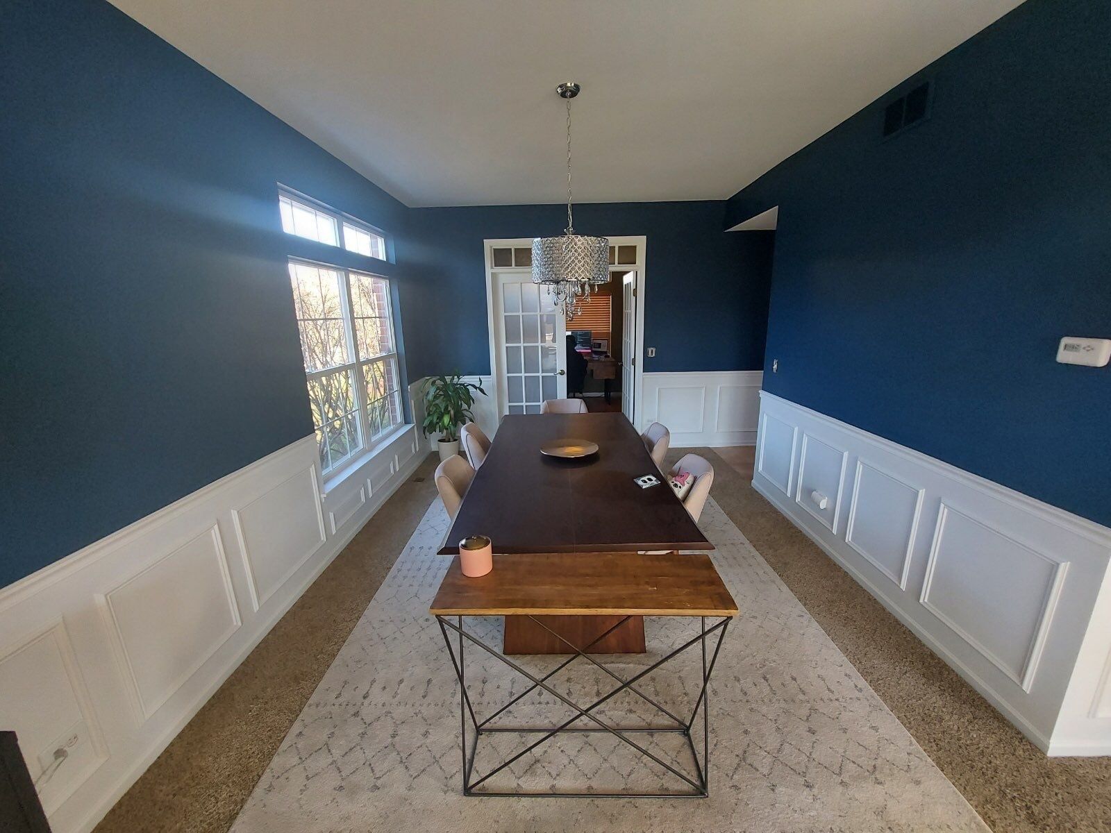 Long dining room with navy blue walls, white wainscoting, and a long table centered by windows.