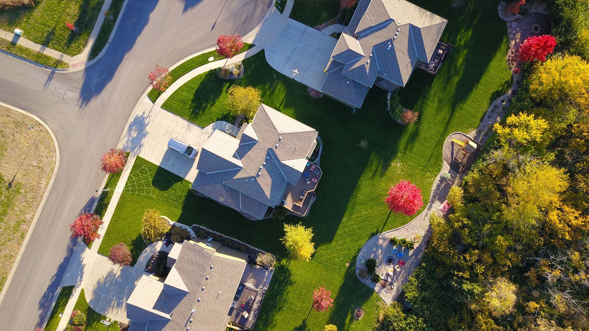 Aerial view of suburban houses with green lawns, trees, and a curved driveway in bright sunlight