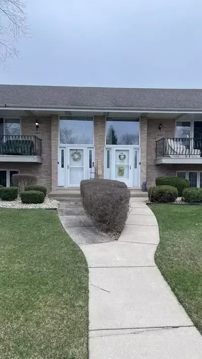 Apartment building entrance with white double doors, brick facade, and a concrete walkway leading past trimmed hedges