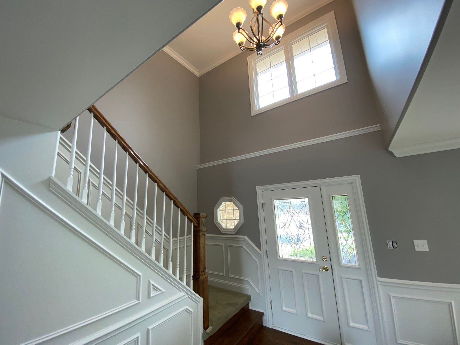 Entry foyer with staircase, white wainscoting, gray walls, and a chandelier under a tall window.