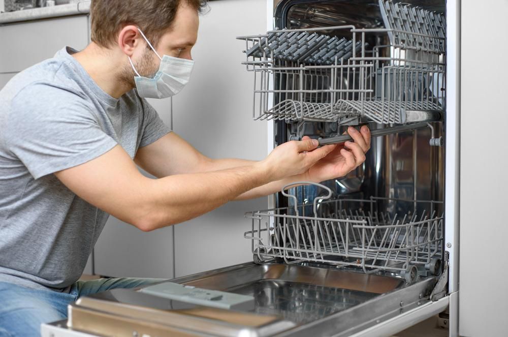 A Man Wearing A Mask Is Fixing A Dishwasher — Gold Coast City Plumbing In Helensvale, QLD