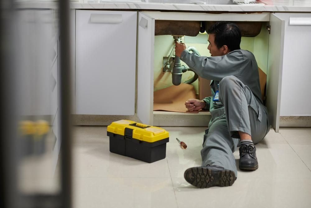 A Man Is Kneeling On The Floor Fixing A Sink In A Kitchen — Gold Coast City Plumbing In Helensvale, QLD