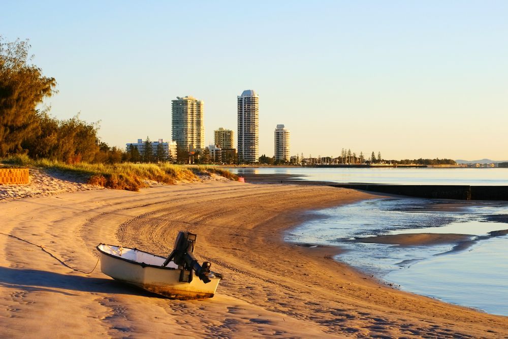 A Small Boat Rests on a Sandy Beach at Sunrise, With High-rise Buildings in the Distance — Gold Coast City Plumbing In Runaway Bay, QLD
