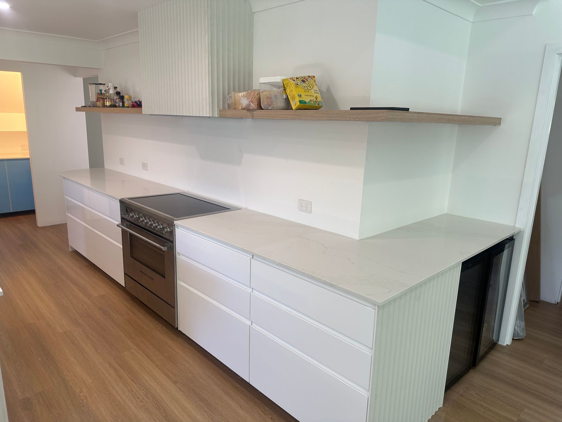 Modern white kitchen with a built-in oven, countertop, and a wooden shelf.