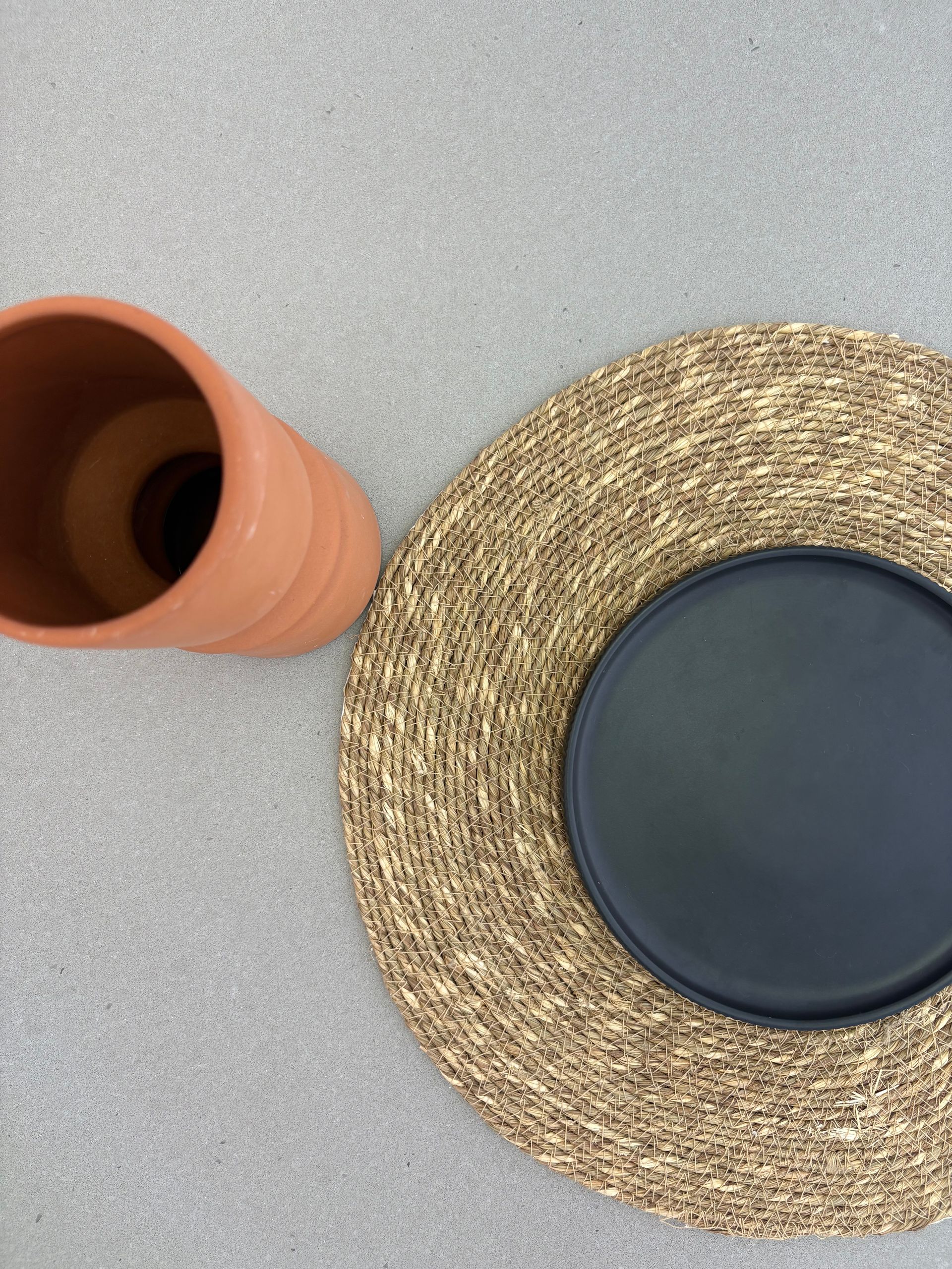 Terracotta vase, black plate on a woven placemat, on a grey surface.