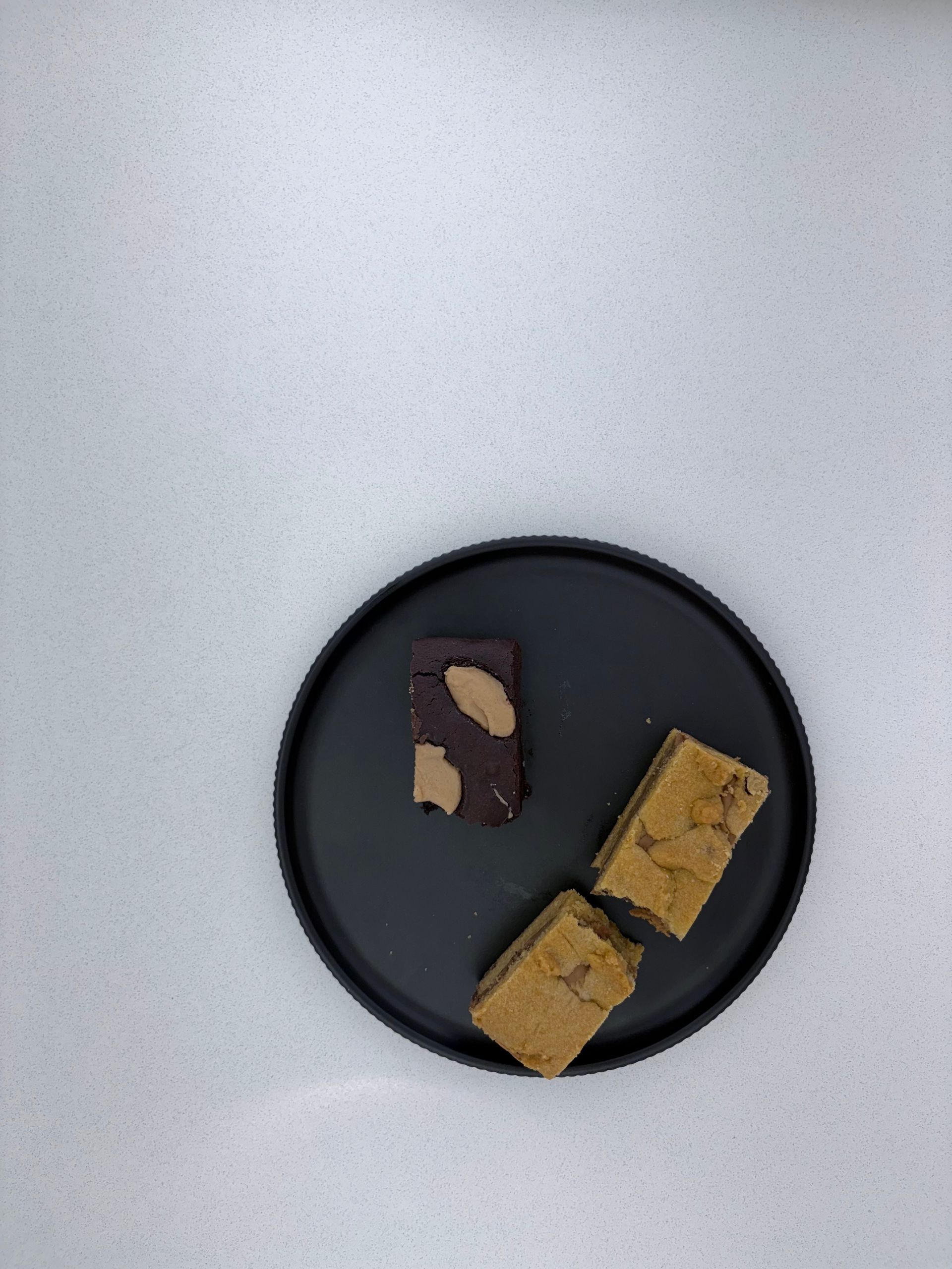 Black plate with three dessert bars, two tan and one brown with almonds, on a white surface.