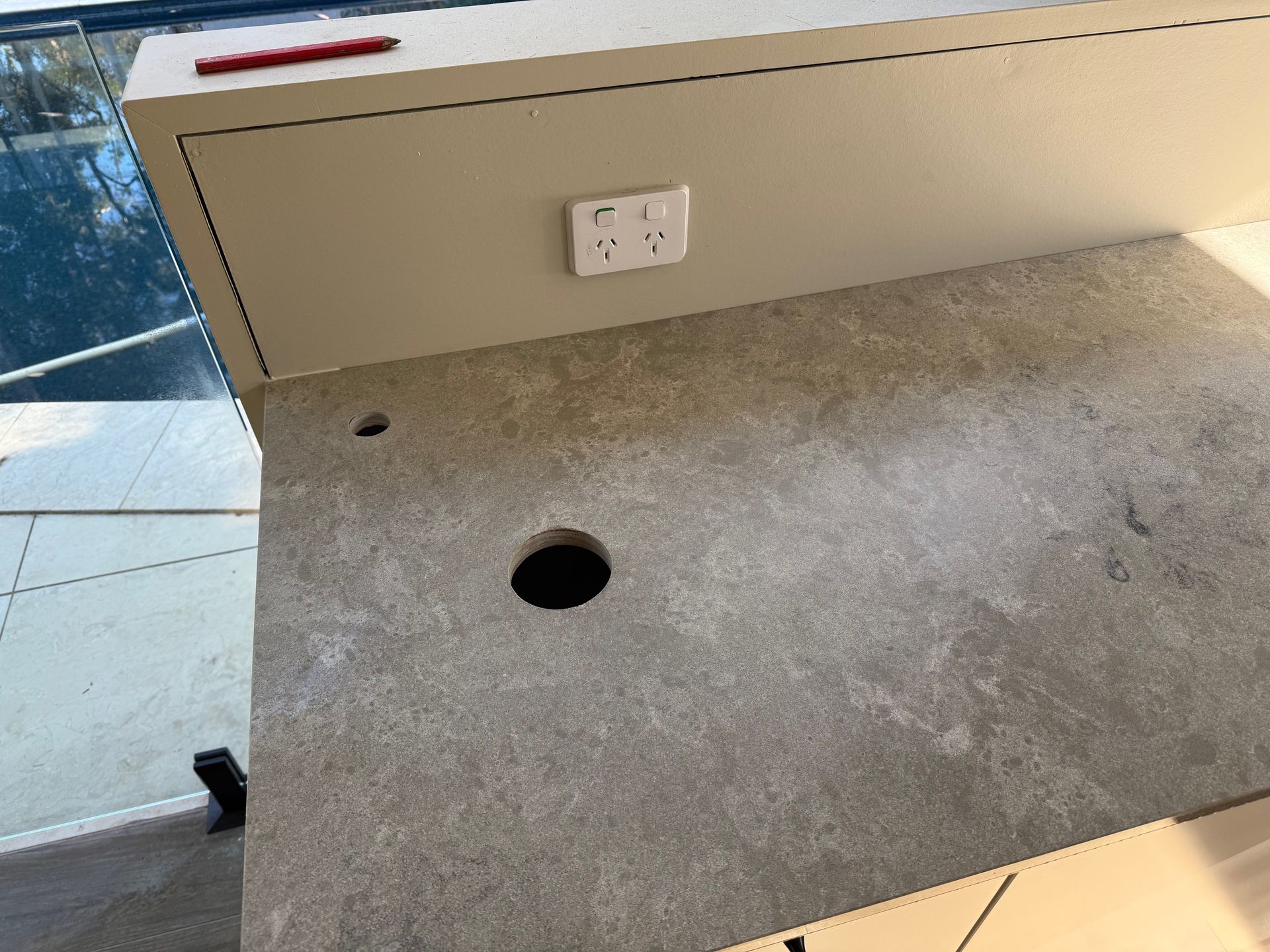 Gray countertop with a large hole, smaller hole, and a power outlet underneath a beige cabinet.