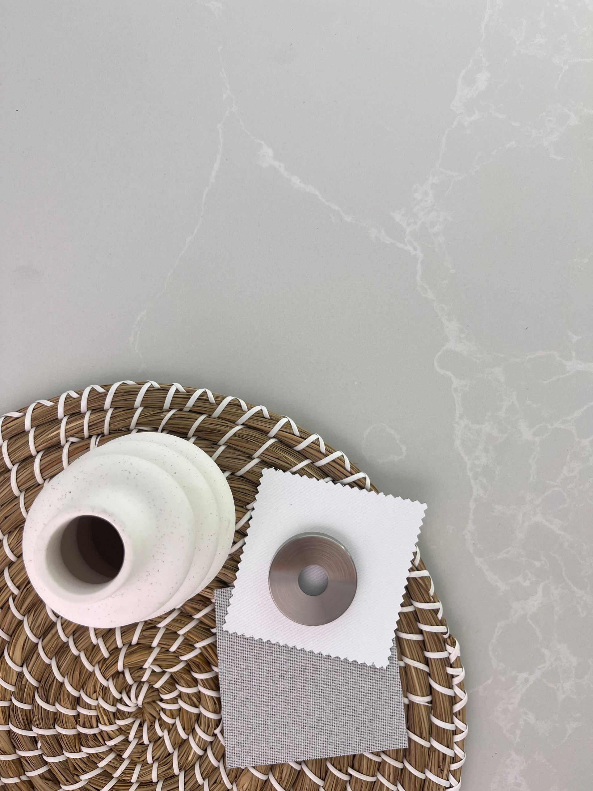 White vase, fabric swatches, and a metal component on a woven placemat, against a marbled gray surface.