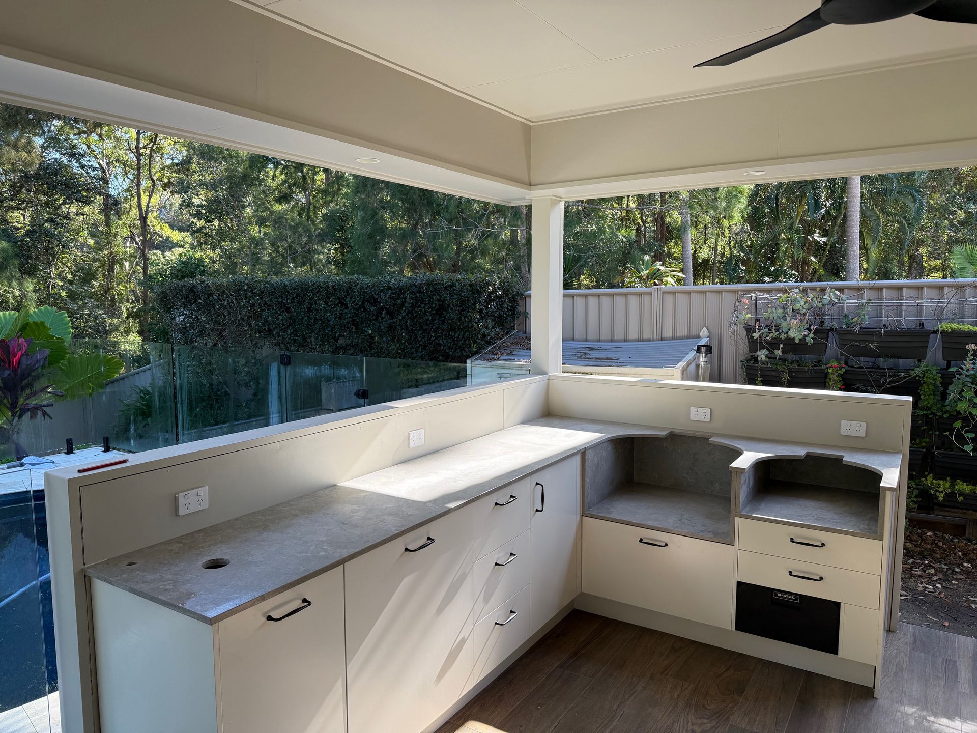 Outdoor kitchen with light cabinets, countertop, and a view of greenery through windows.