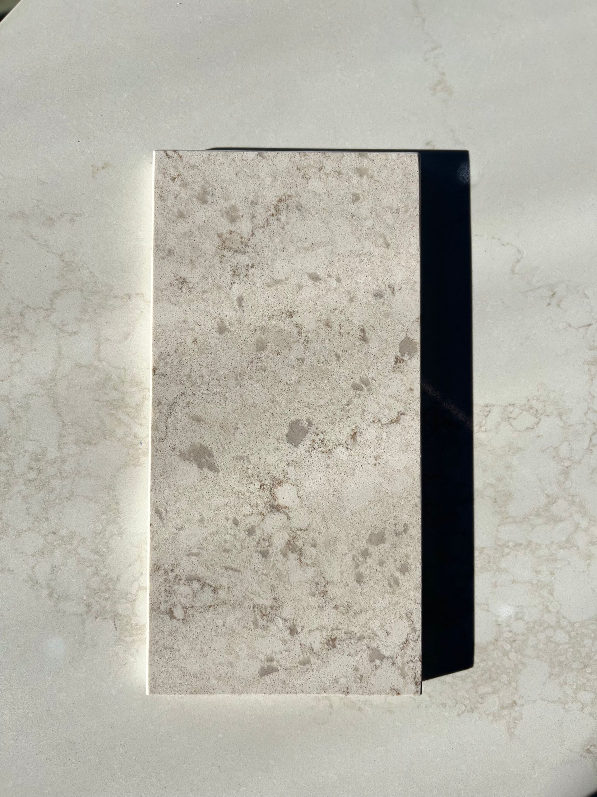 Rectangular beige stone tile with speckled pattern, casting a dark shadow on a light surface.
