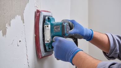 Person sanding a wall with a power sander, wearing blue gloves and a dust mask