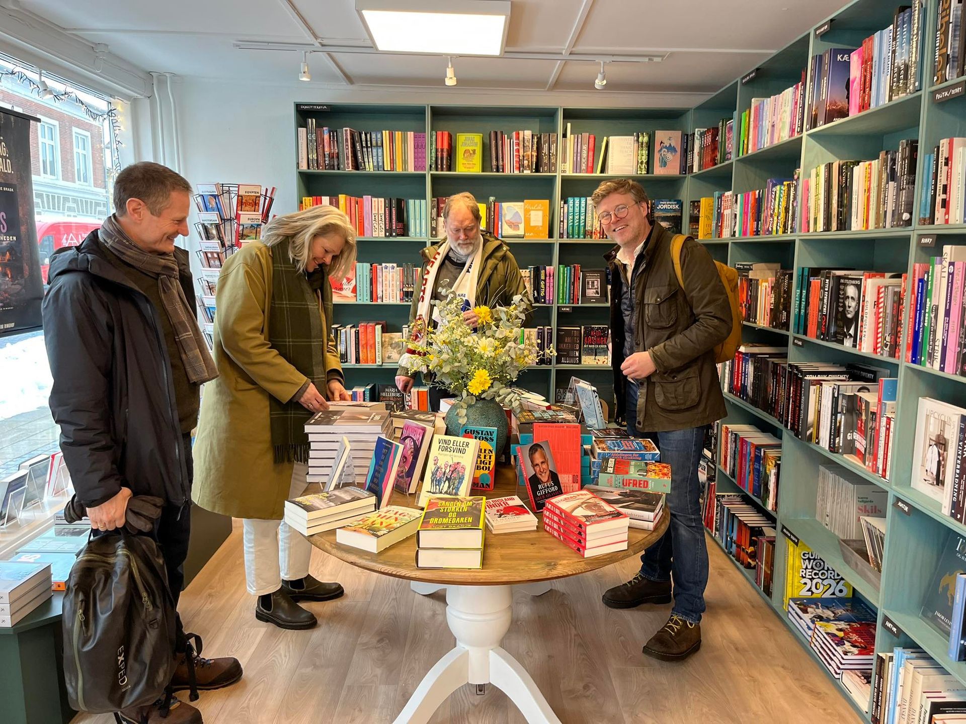 Folk kigger i bøger og interagerer omkring et rundt udstillingsbord i en klart oplyst, hyggelig boghandel.