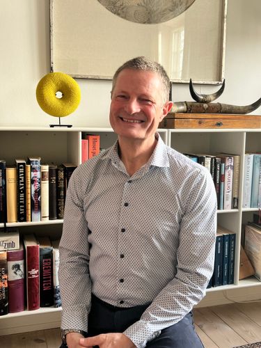 En smilende mand i en mønstret skjorte med knapper sidder foran en bogreol med bøger og dekorative kunstgenstande.
