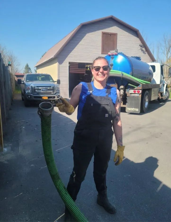 Smiling worker in overalls holding a green hose beside a blue tank truck in a driveway