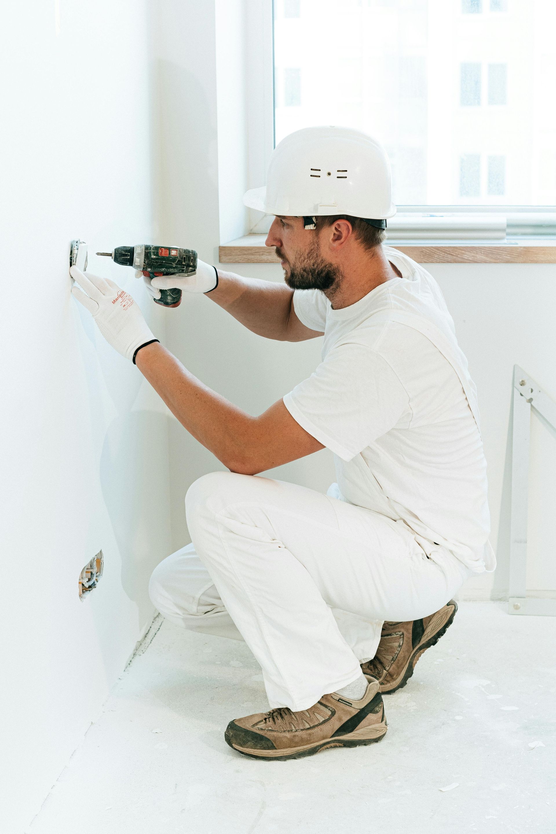 Man in white work clothes and hard hat using a drill on a wall.