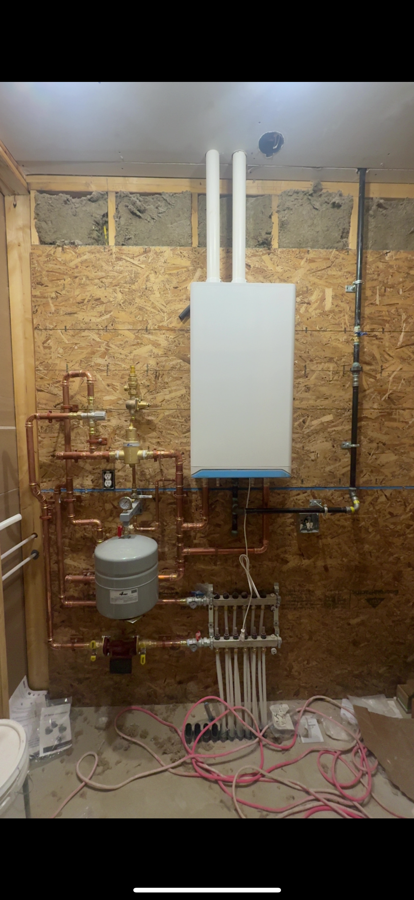 Utility room with a white boiler, exposed pipes, wires, and unfinished plywood walls.