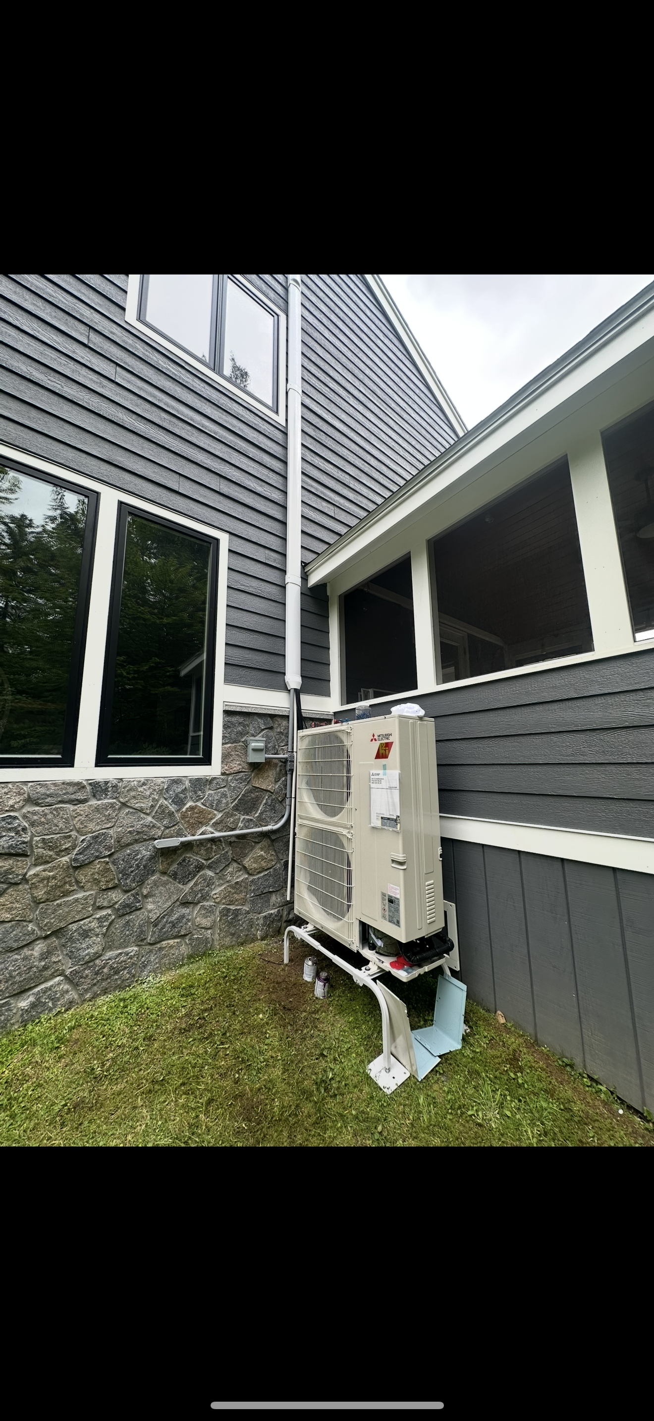 Outdoor HVAC unit beside a gray building with stone siding and a small grass strip