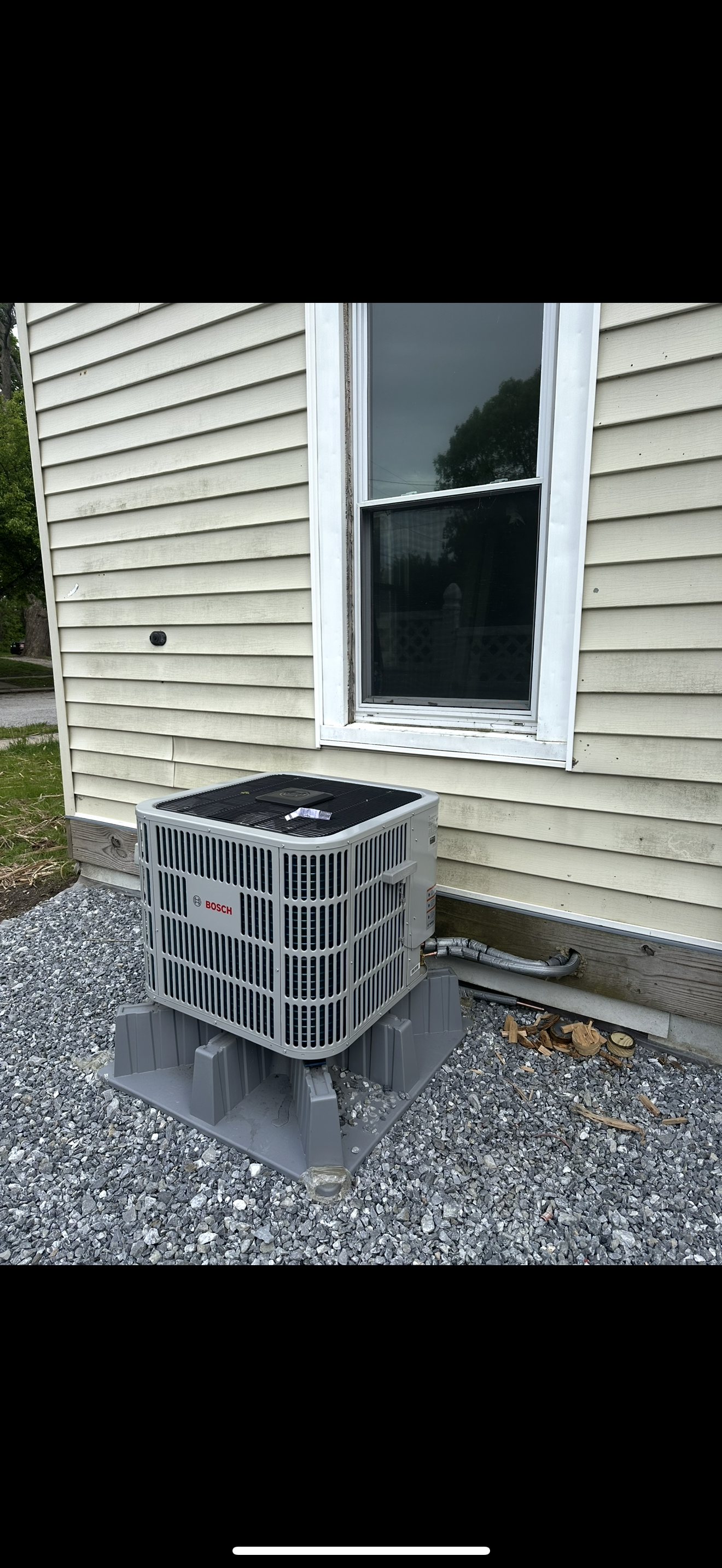 Outdoor air conditioner unit beside a beige house wall under a window, sitting on gravel