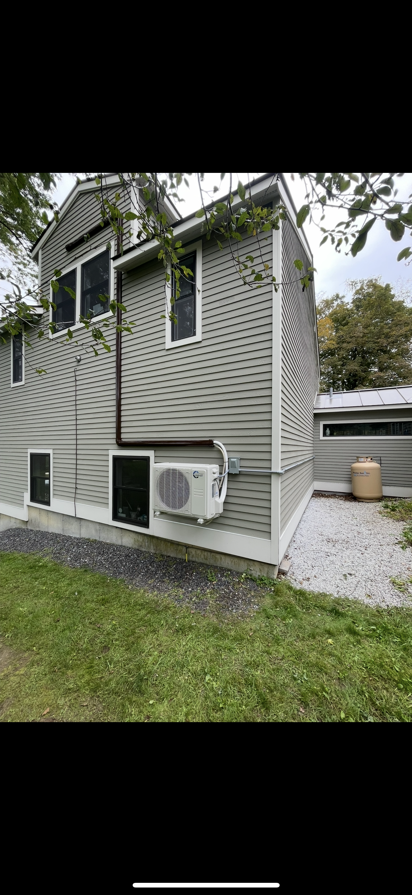 Small gray house with white trim, windows, and an air conditioner beside a gravel driveway and green lawn.