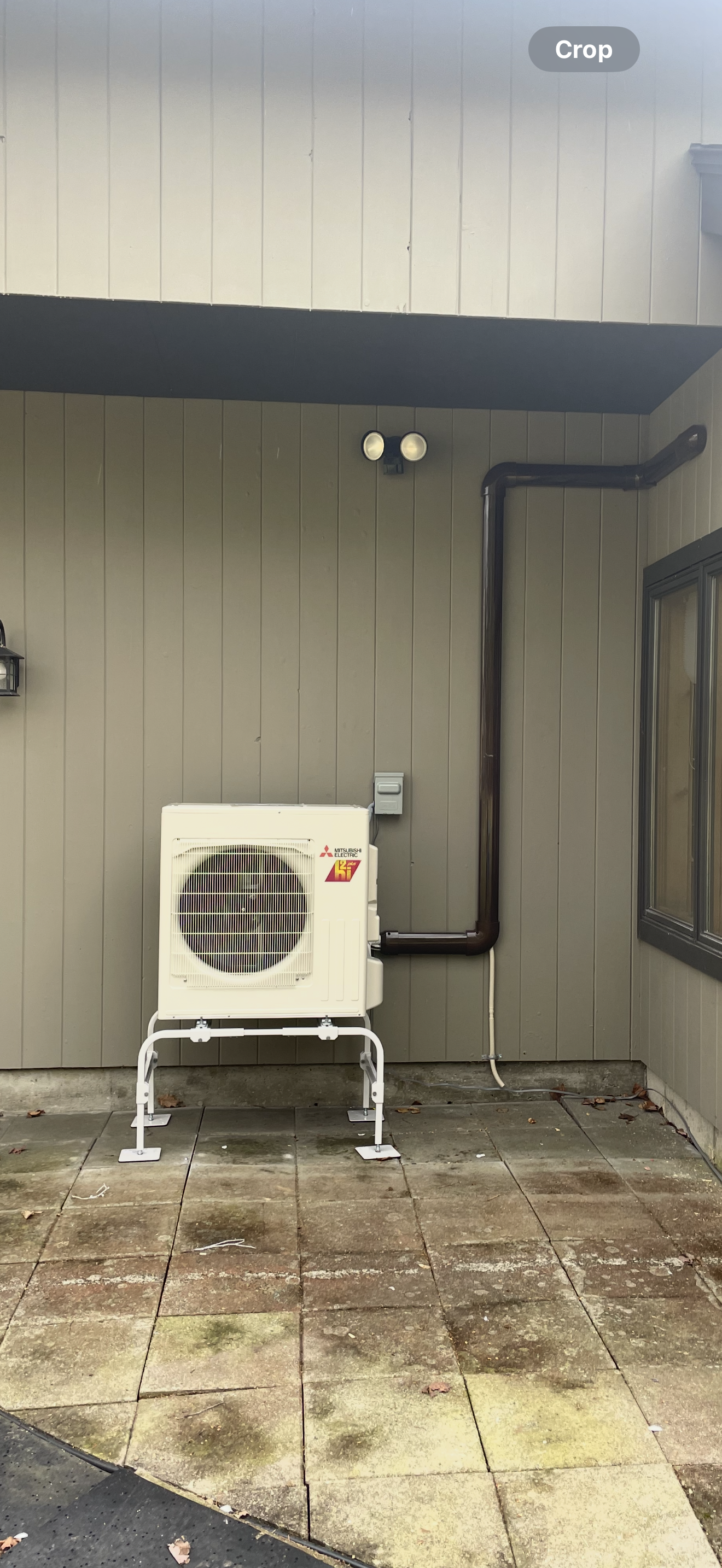 Outdoor HVAC unit beside a gray house wall under a dark awning, with pipes and damp concrete below