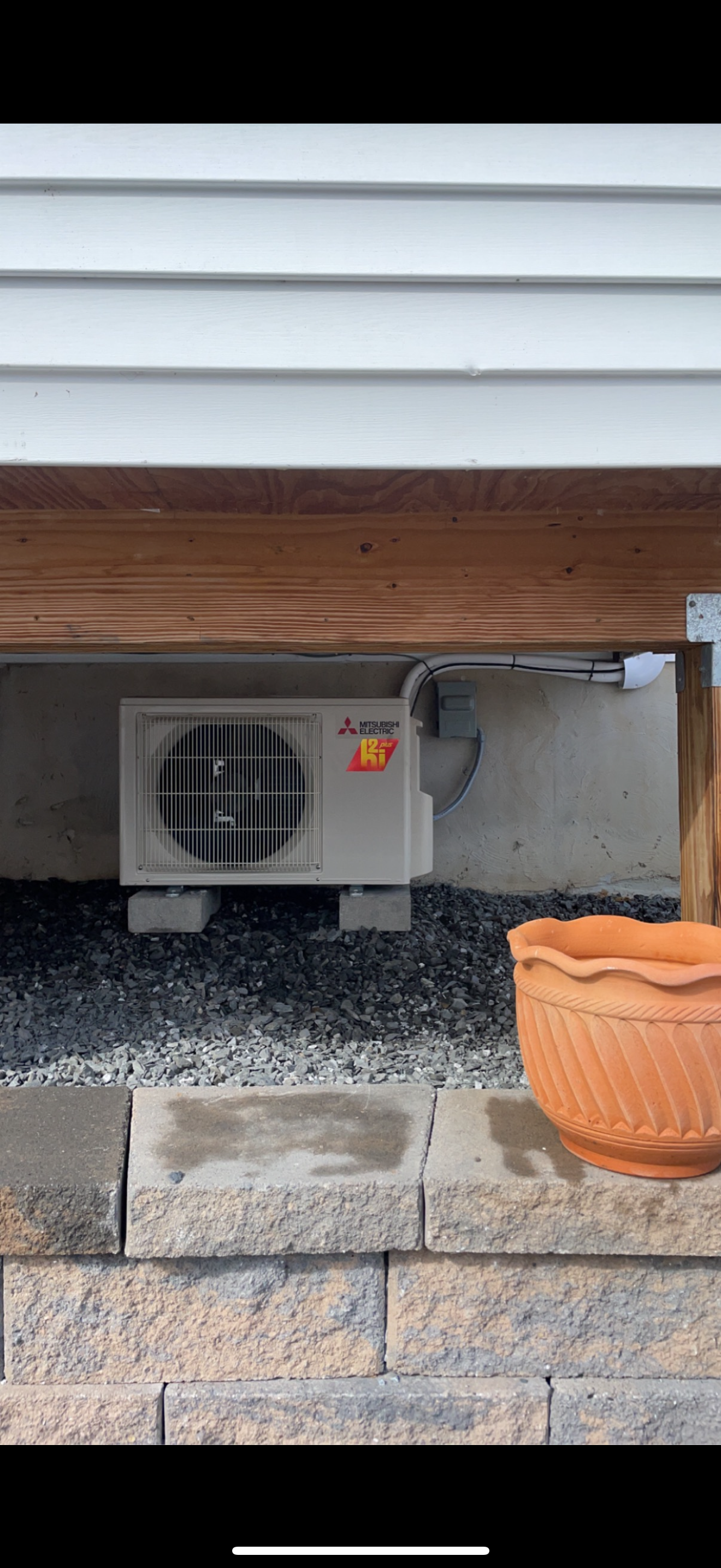 Outdoor HVAC unit beneath a deck, beside gray gravel, stone steps, and a terracotta planter.