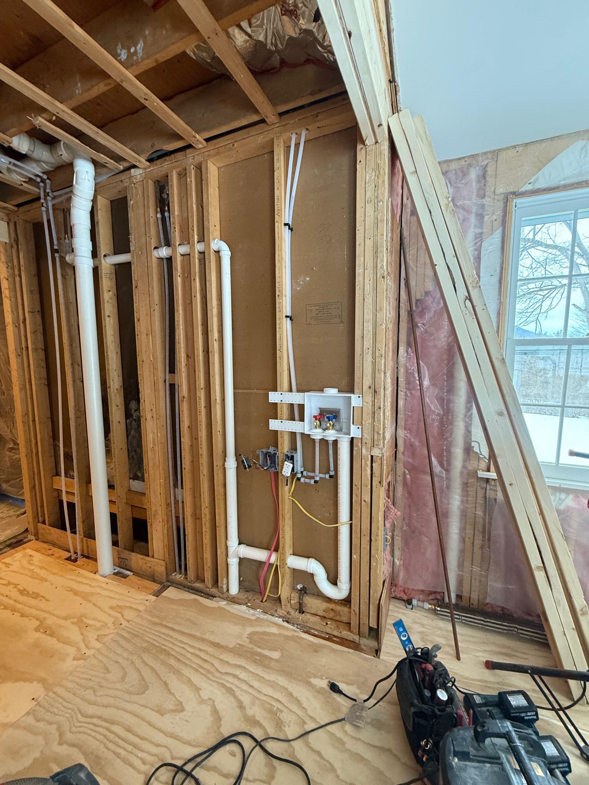 Interior wall under renovation with exposed studs, plumbing, and window light on a plywood floor