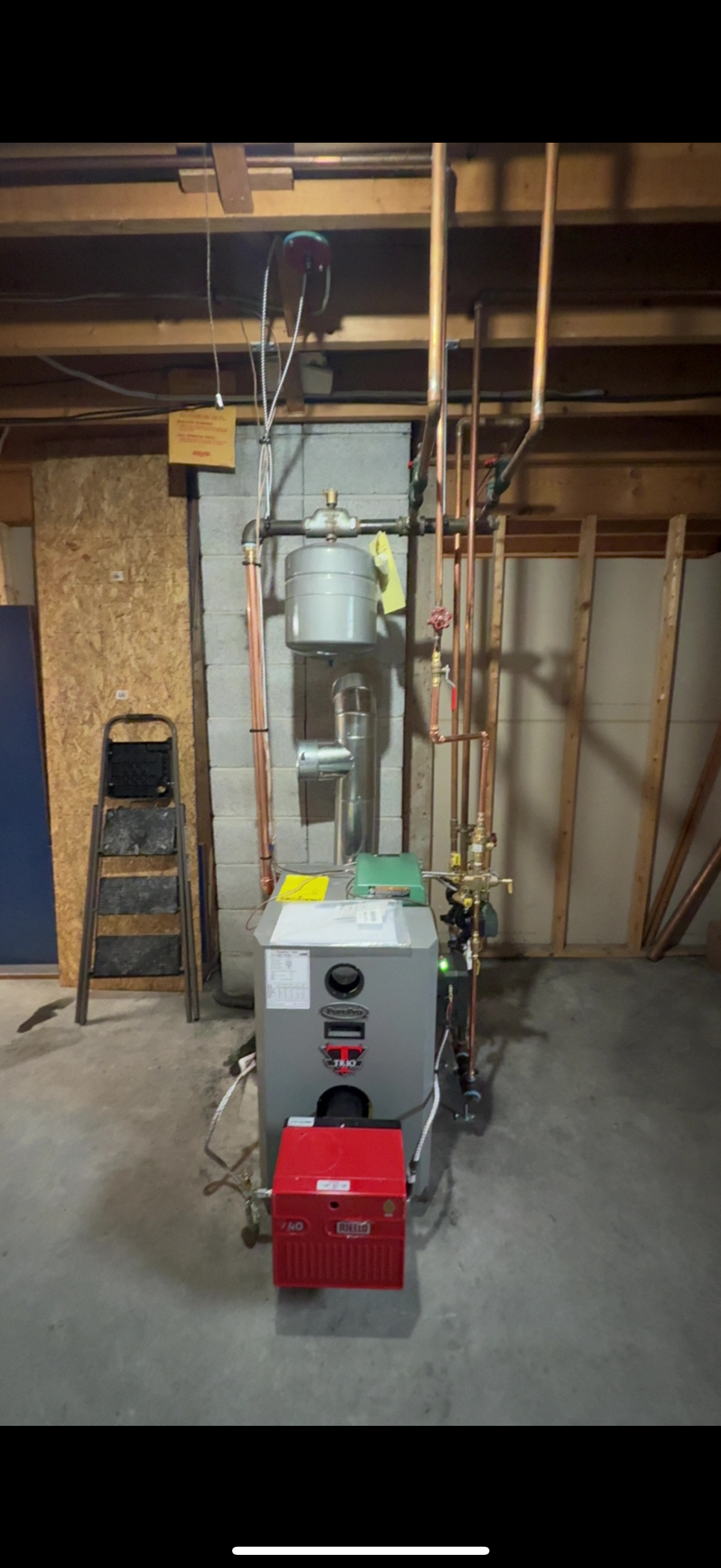 Boiler and water heater in an unfinished basement, with exposed pipes and wood framing.