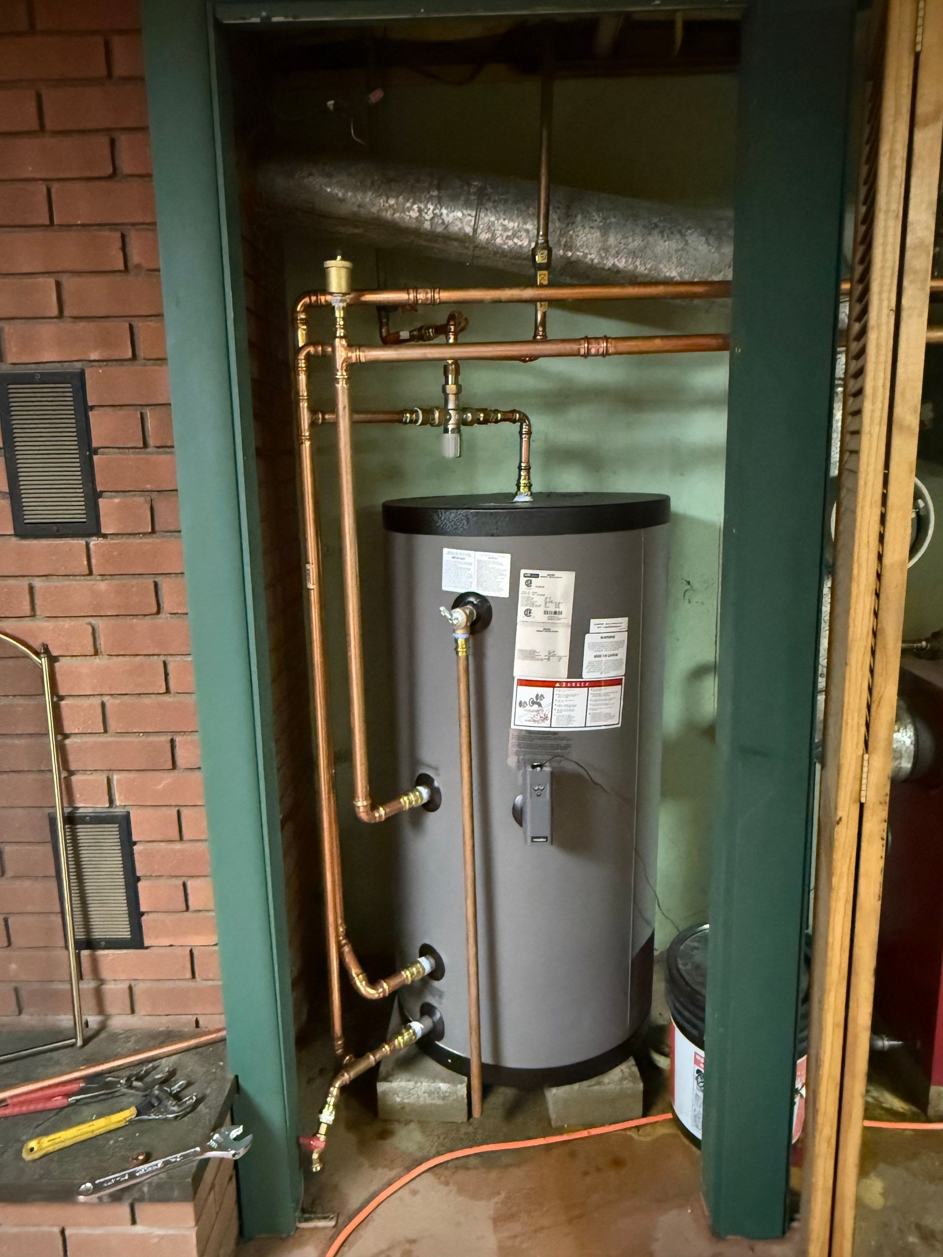 Water heater in a narrow utility closet with copper pipes and exposed brick walls.