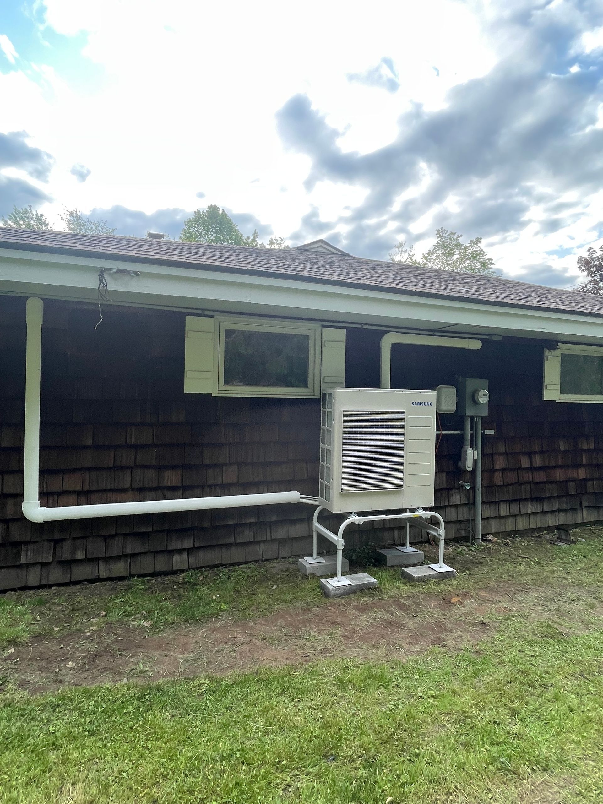 Old shingle building with a window air conditioner under a cloudy sky, beside a grassy yard